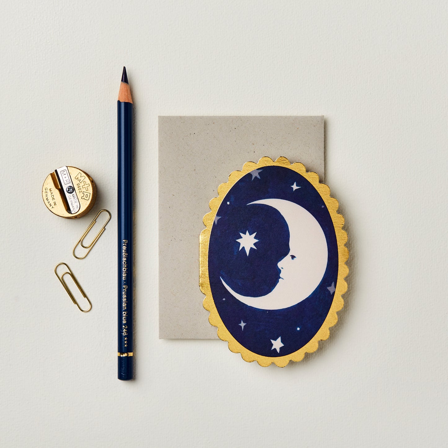 A navy blue card featuring a crescent moon and star design sits on a light background. Beside it, a sharpened pencil, a gold pencil sharpener, and two paperclips are arranged neatly, creating a serene and artistic workspace.