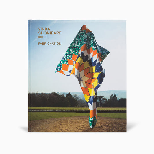 A book cover featuring the title "Yinka Shonibare MBE: Fabric-ation" with a vibrant, patterned sculpture resembling a flag or fabric in a landscape setting. The background shows a clear sky and greenery, highlighting the artwork's bold colours.