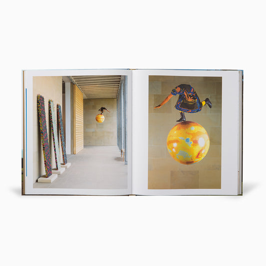 A photo book open to two pages. On the left, colourful vertical sculptures stand against a wall. On the right, a figure in traditional attire balances on a large, vibrant yellow sphere, set against a minimalist background.