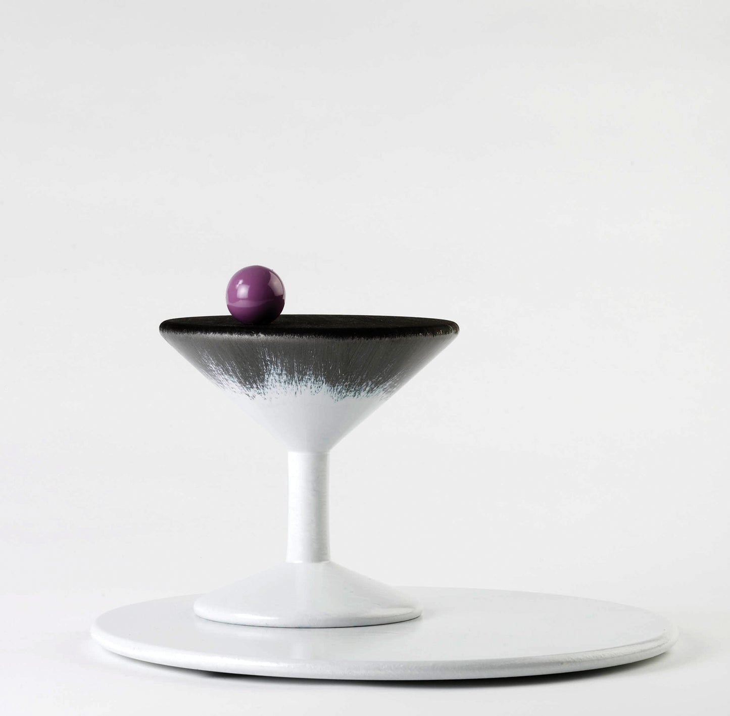 A minimalist 3D cocktail glass with a black and white design sits on a white plate. A single purple olive rests on the rim of the glass, creating a striking contrast against the clean background.