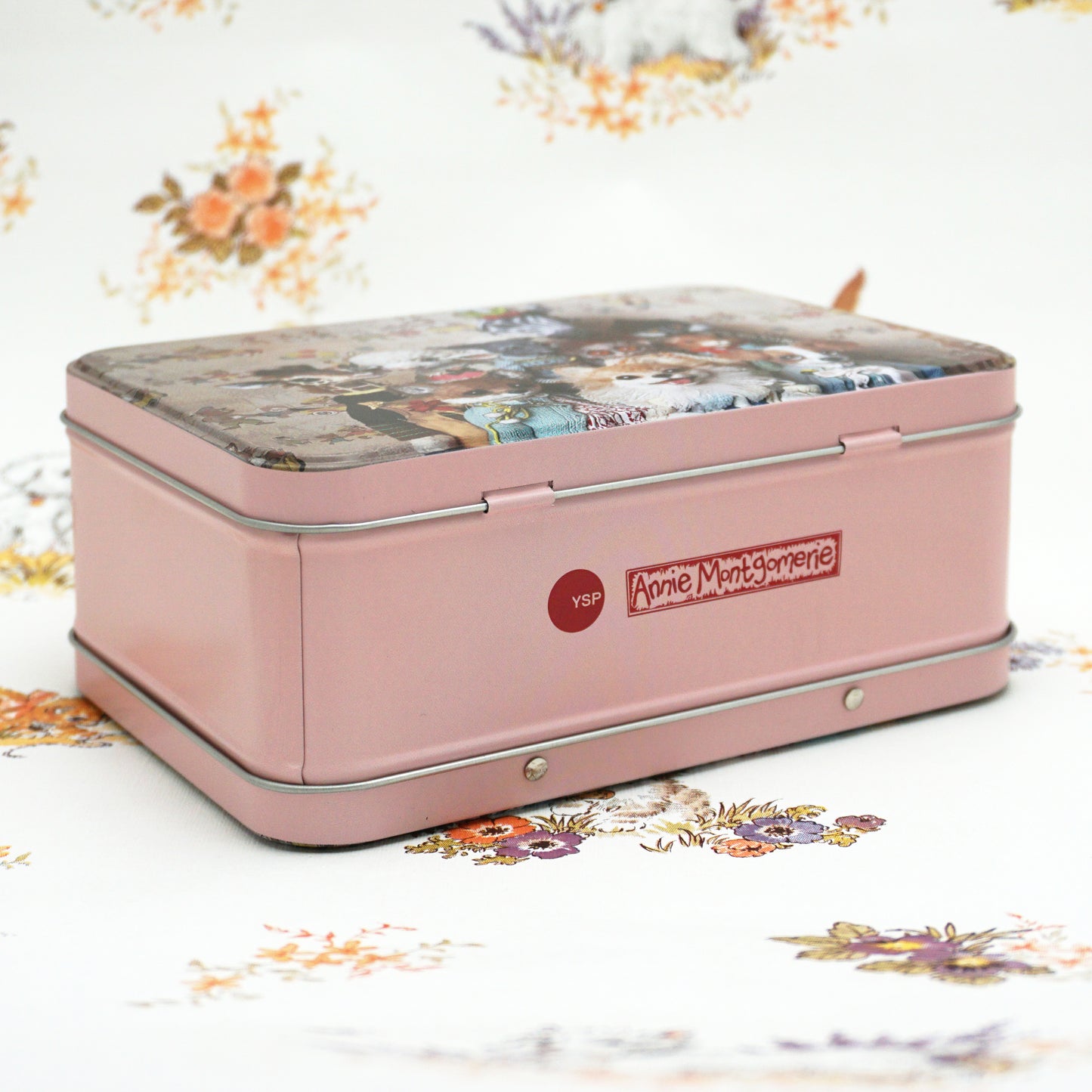 A vintage-style pink tin box featuring a decorative lid with a floral and pastoral scene. The box is labelled "Annie Montgomerie" and has a shiny silver trim, resting on a patterned floral background.