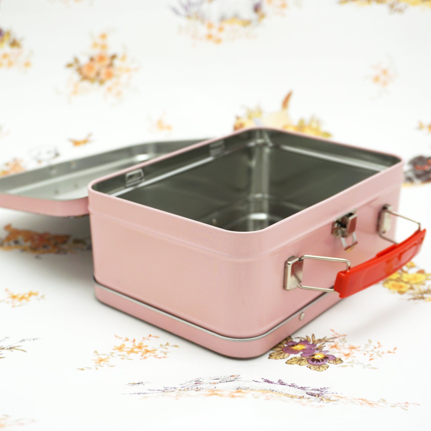 A pink metal lunchbox with a shiny silver interior is shown open, revealing its rectangular shape. The box features a bright red handle and a metal clasp on the front, set against a floral patterned background.
