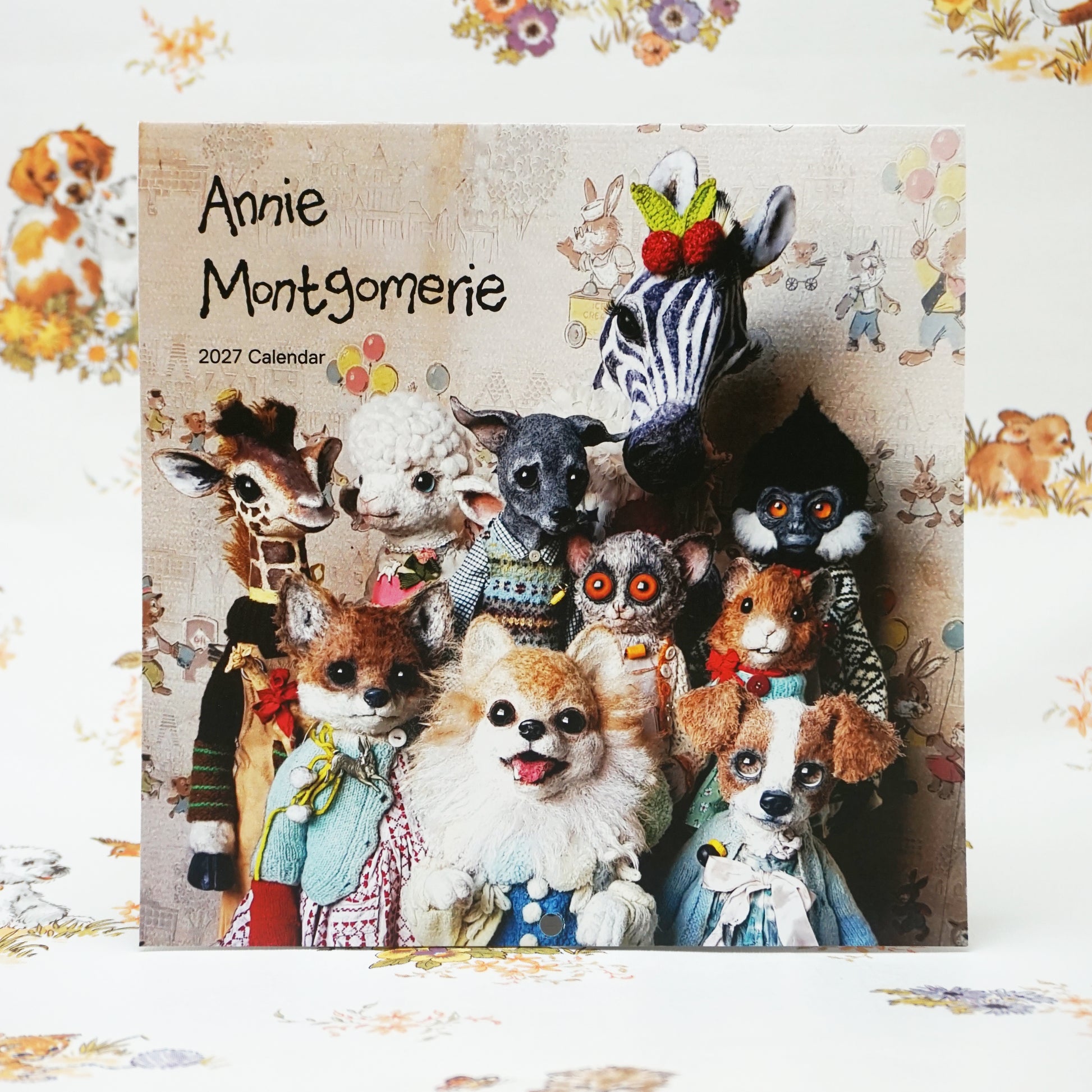 A calendar cover featuring a group of whimsical, plush animal figures, including a zebra, giraffe, fox, and dog, all dressed in colourful clothing. The text reads "Annie Montgomerie 2027 Calendar" against a patterned background.