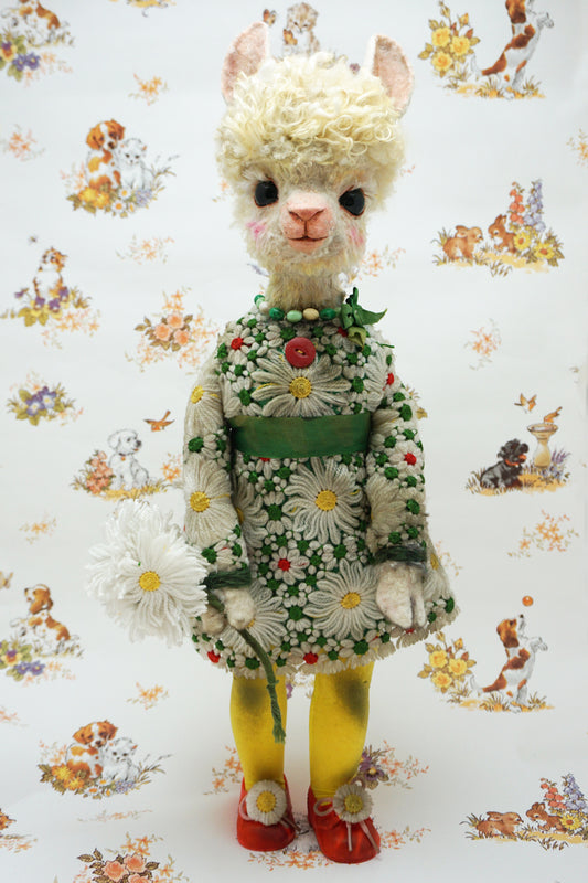 A handmade plush alpaca doll with curly white hair, wearing a green and white daisy-patterned dress, yellow tights, and red shoes, holding a white daisy bouquet against a patterned background.
