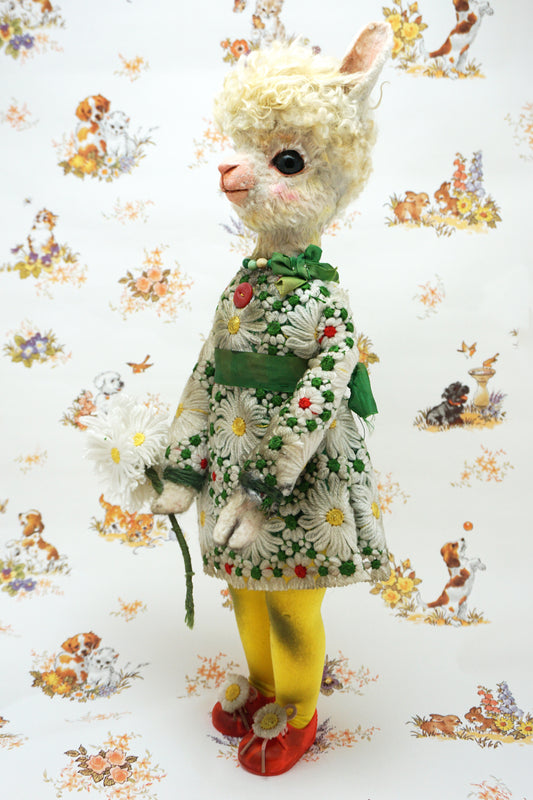 A vintage-style plush alpaca doll wearing a white daisy-patterned dress with green trim, yellow tights, and red shoes, standing against a white background with a repeating printed pattern of small animals.