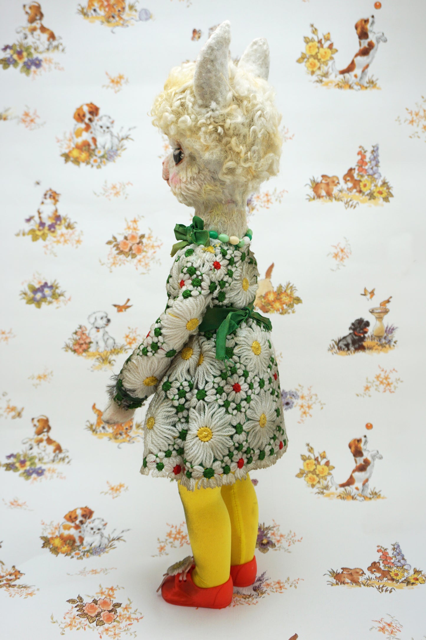 A vintage-style plush alpaca doll shown from the back, wearing a white floral dress with a green bow, yellow tights, and red shoes, standing against a white patterned backdrop.