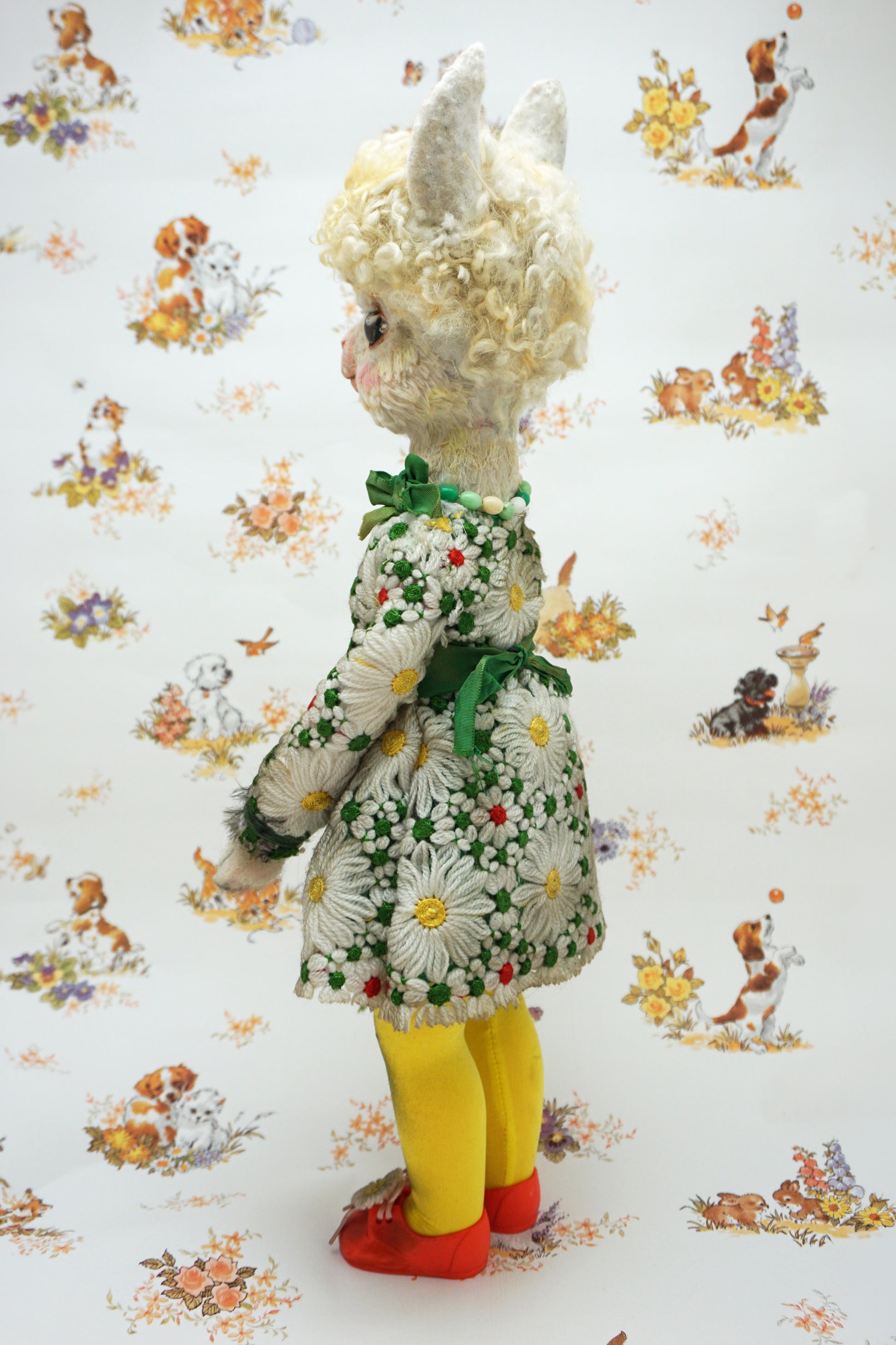A vintage-style plush alpaca doll shown from the back, wearing a white floral dress with a green bow, yellow tights, and red shoes, standing against a white patterned backdrop.