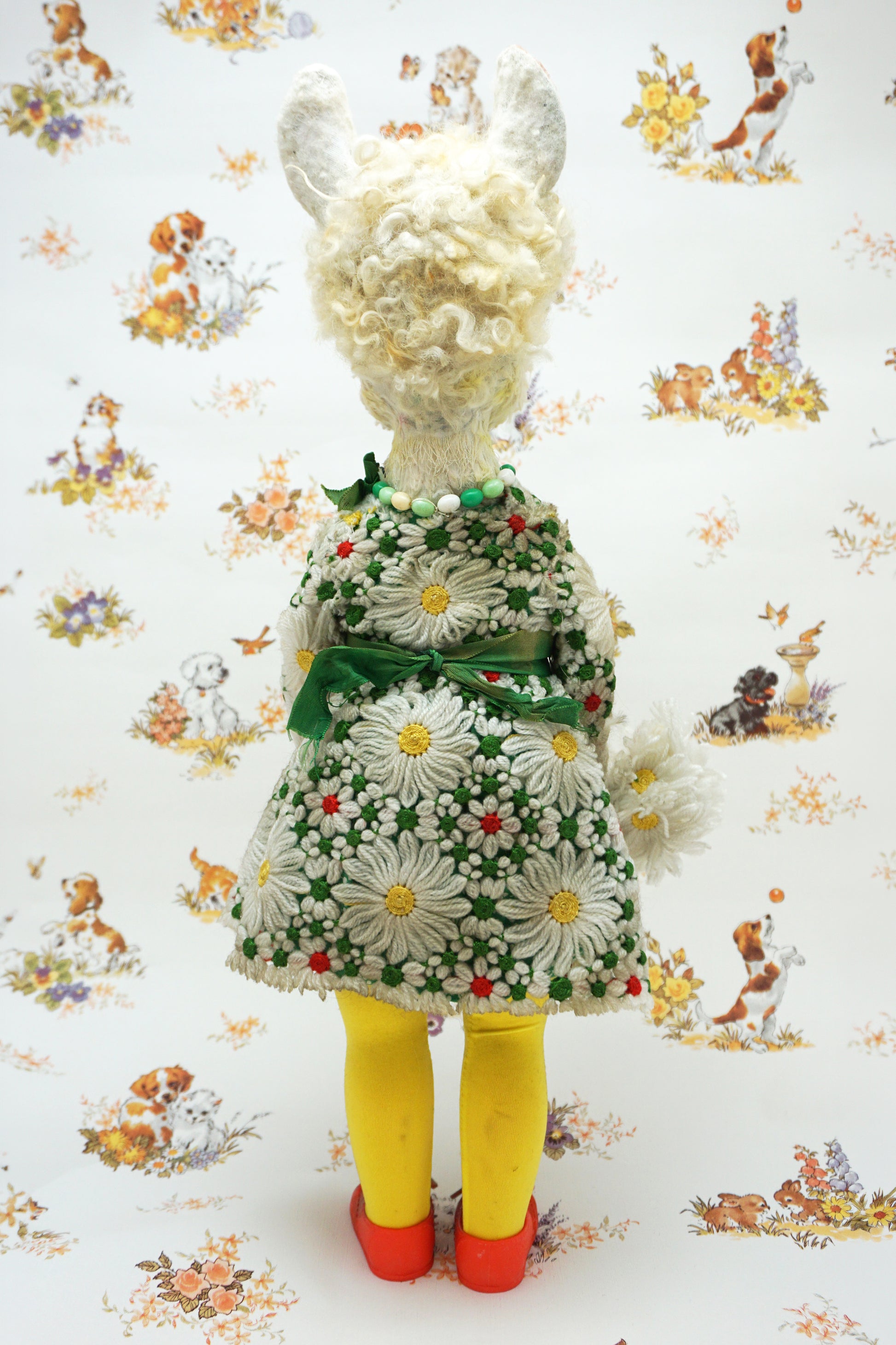 A vintage-style doll viewed from the back, wearing a green and white daisy-patterned dress with a green ribbon, yellow tights, and red shoes, standing against a white background with a matching daisy and animal print.