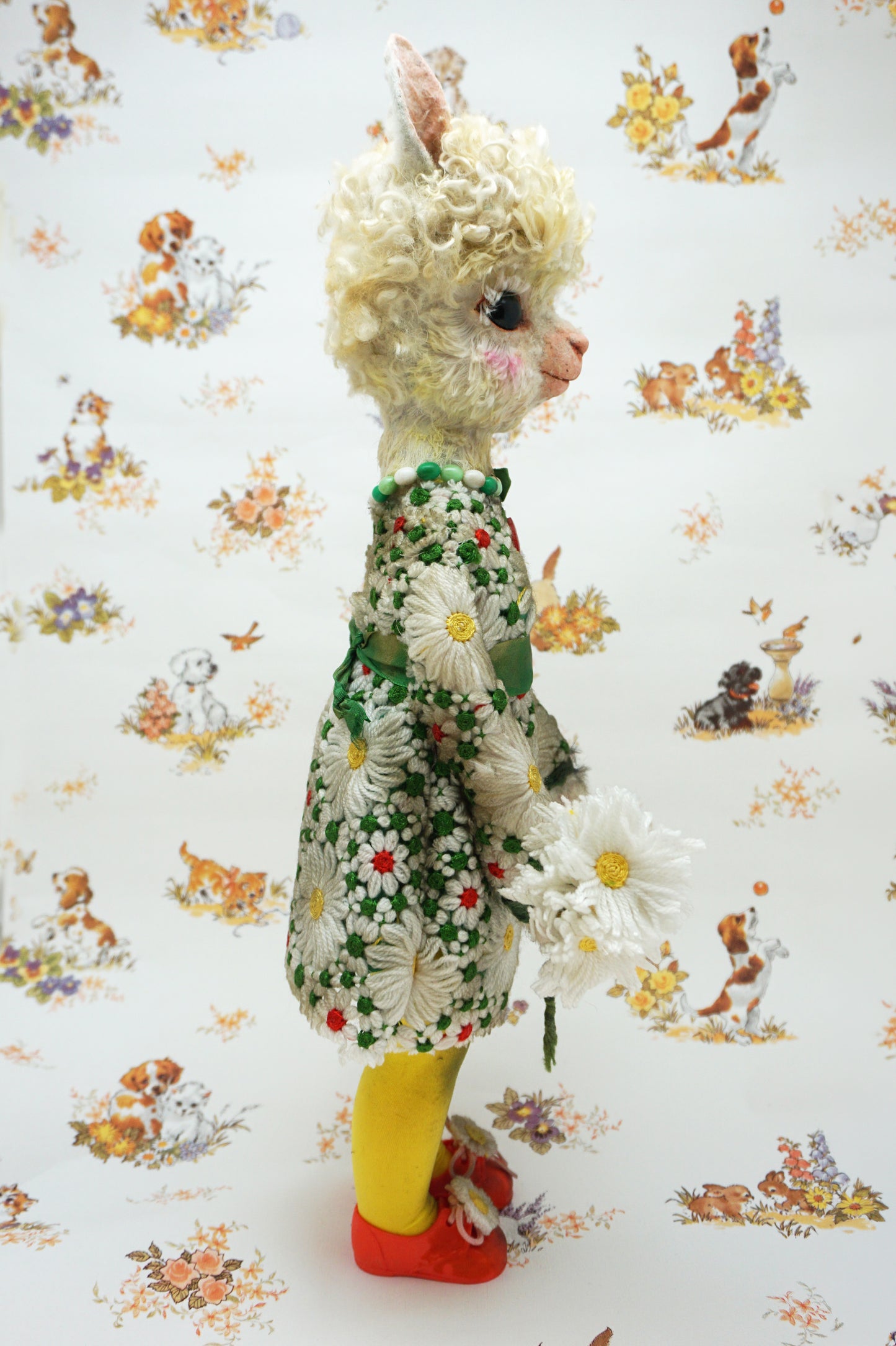 A vintage-style plush alpaca doll with curly blonde hair, wearing a green and white daisy-patterned dress, yellow tights, and red shoes, posed against a white background with a matching daisy-and-animal patterned paper backdrop.