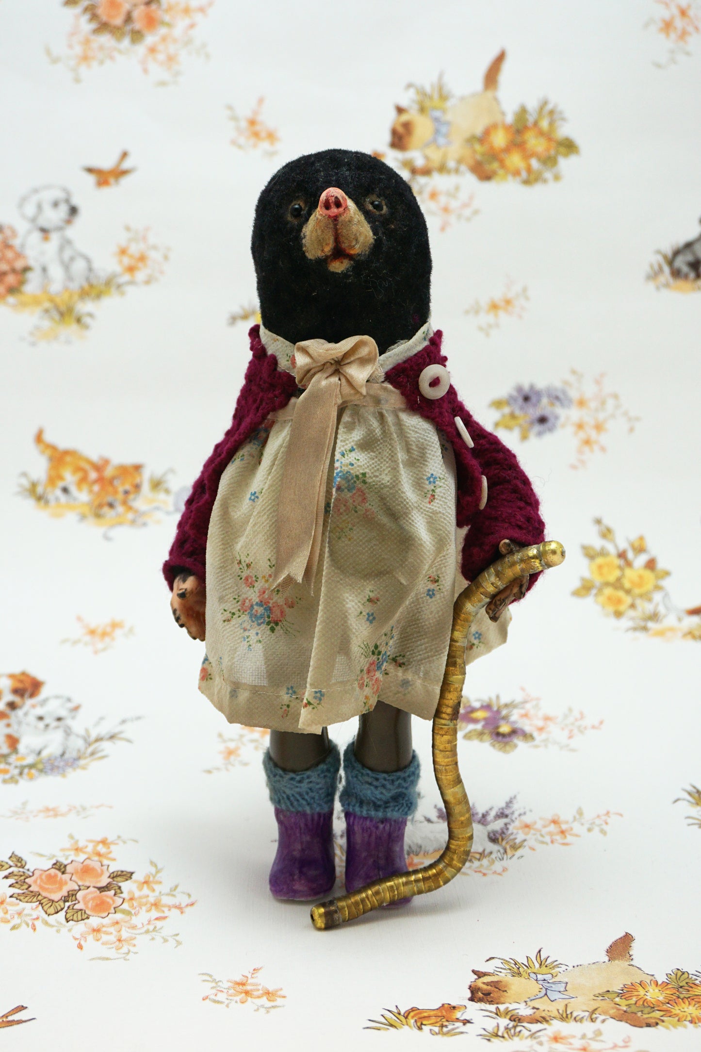 A handmade mole, dressed in a burgundy cardigan, a cream floral dress with a beige ribbon bow, and purple knit socks, holding a curved gold-coloured worm. The toy is posed against a white background with a vintage-style floral and animal print.
