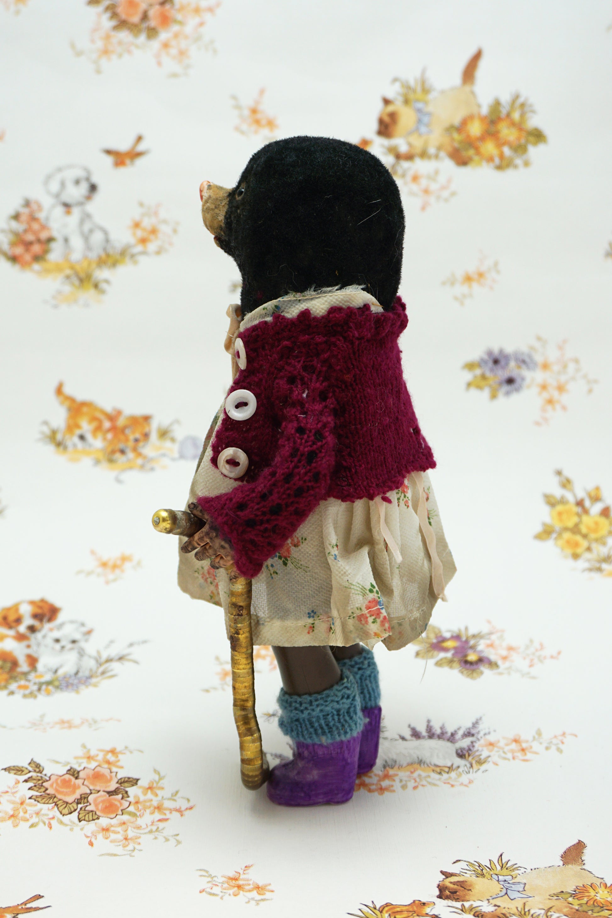 A handmade mole, dressed in a burgundy cardigan, a cream floral dress with a beige ribbon bow, and purple knit socks, holding a curved gold-coloured worm. The toy is posed against a white background with a vintage-style floral and animal print.