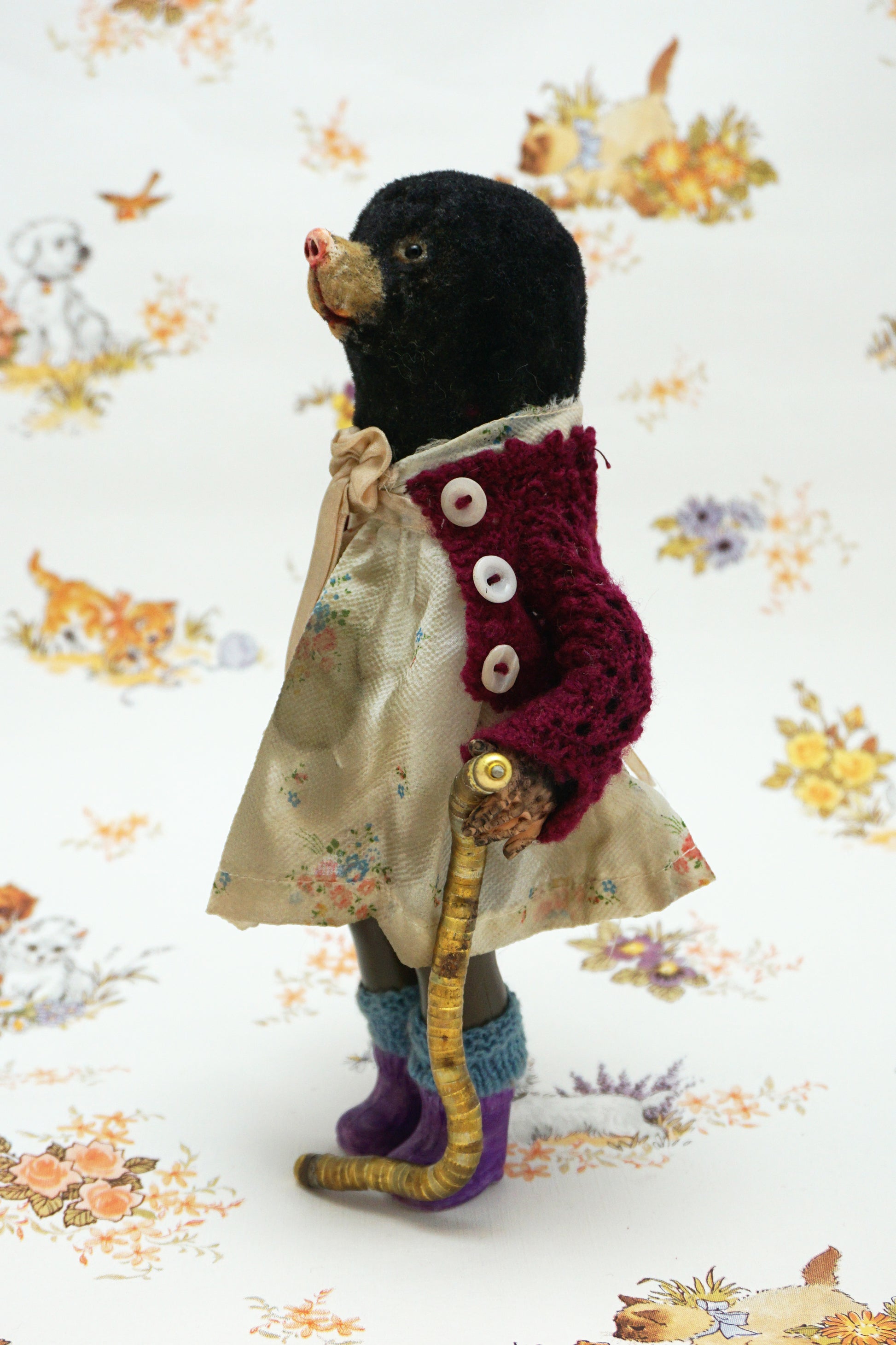 A handmade mole, dressed in a burgundy cardigan, a cream floral dress with a beige ribbon bow, and purple knit socks, holding a curved gold-coloured worm. The toy is posed against a white background with a vintage-style floral and animal print.