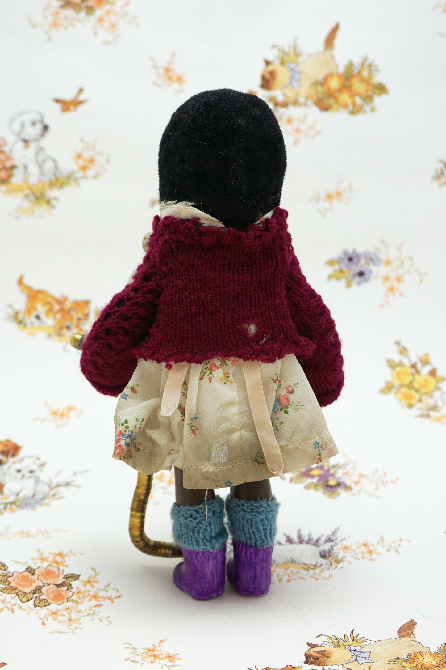 A handmade mole, dressed in a burgundy cardigan, a cream floral dress with a beige ribbon bow, and purple knit socks, holding a curved gold-coloured worm. The toy is posed against a white background with a vintage-style floral and animal print.