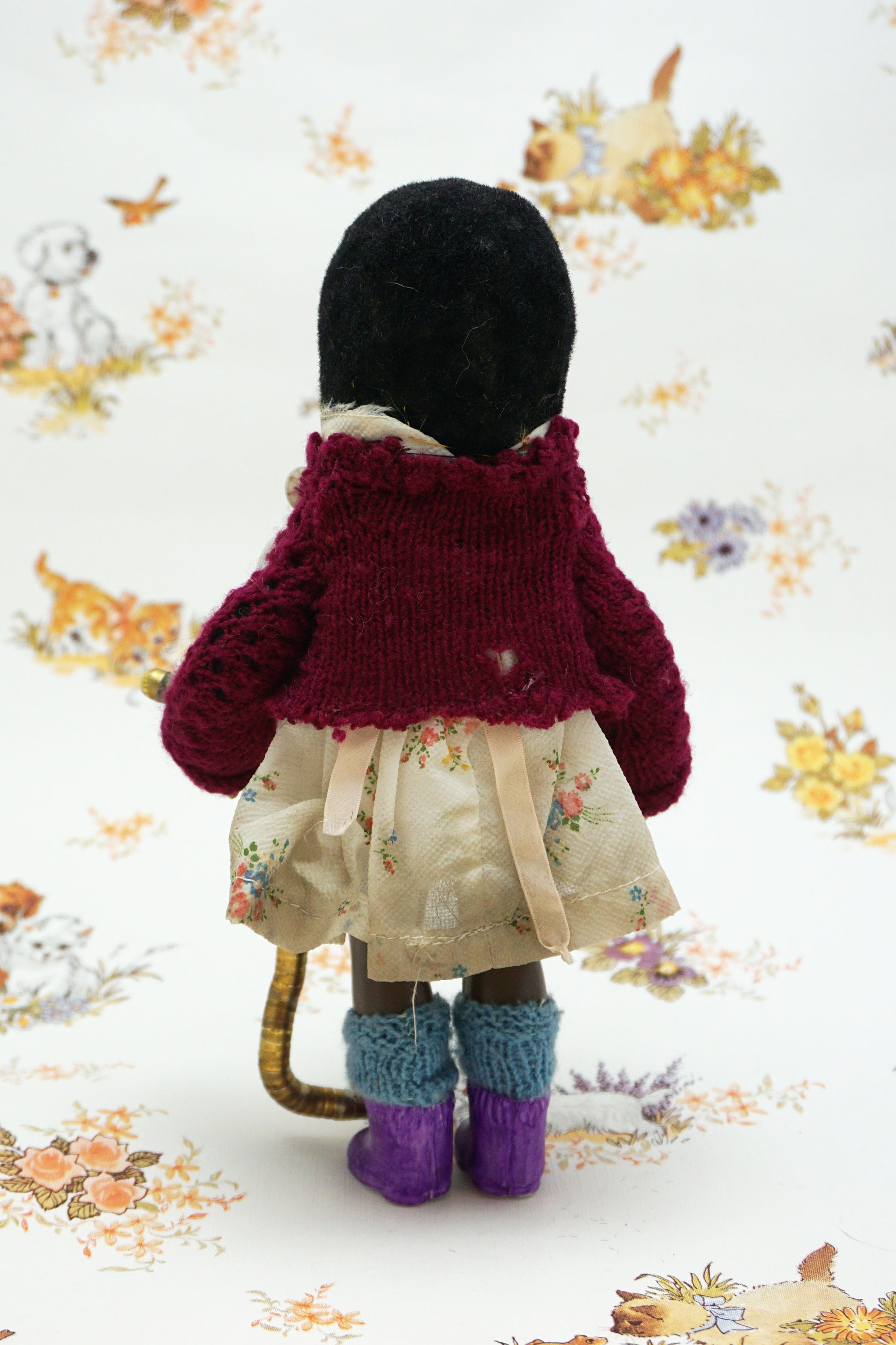 A handmade mole, dressed in a burgundy cardigan, a cream floral dress with a beige ribbon bow, and purple knit socks, holding a curved gold-coloured worm. The toy is posed against a white background with a vintage-style floral and animal print.