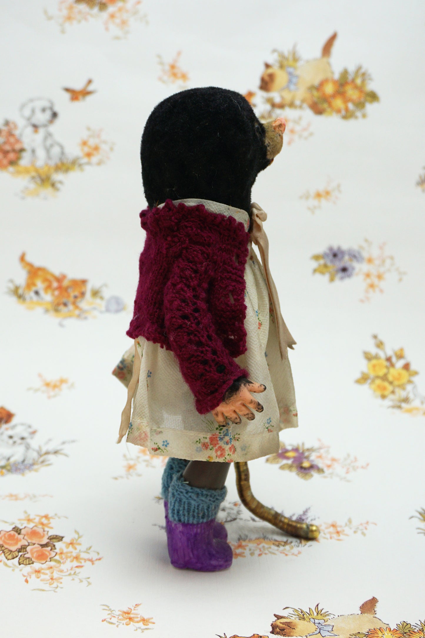 A handmade mole, dressed in a burgundy cardigan, a cream floral dress with a beige ribbon bow, and purple knit socks, holding a curved gold-coloured worm. The toy is posed against a white background with a vintage-style floral and animal print.