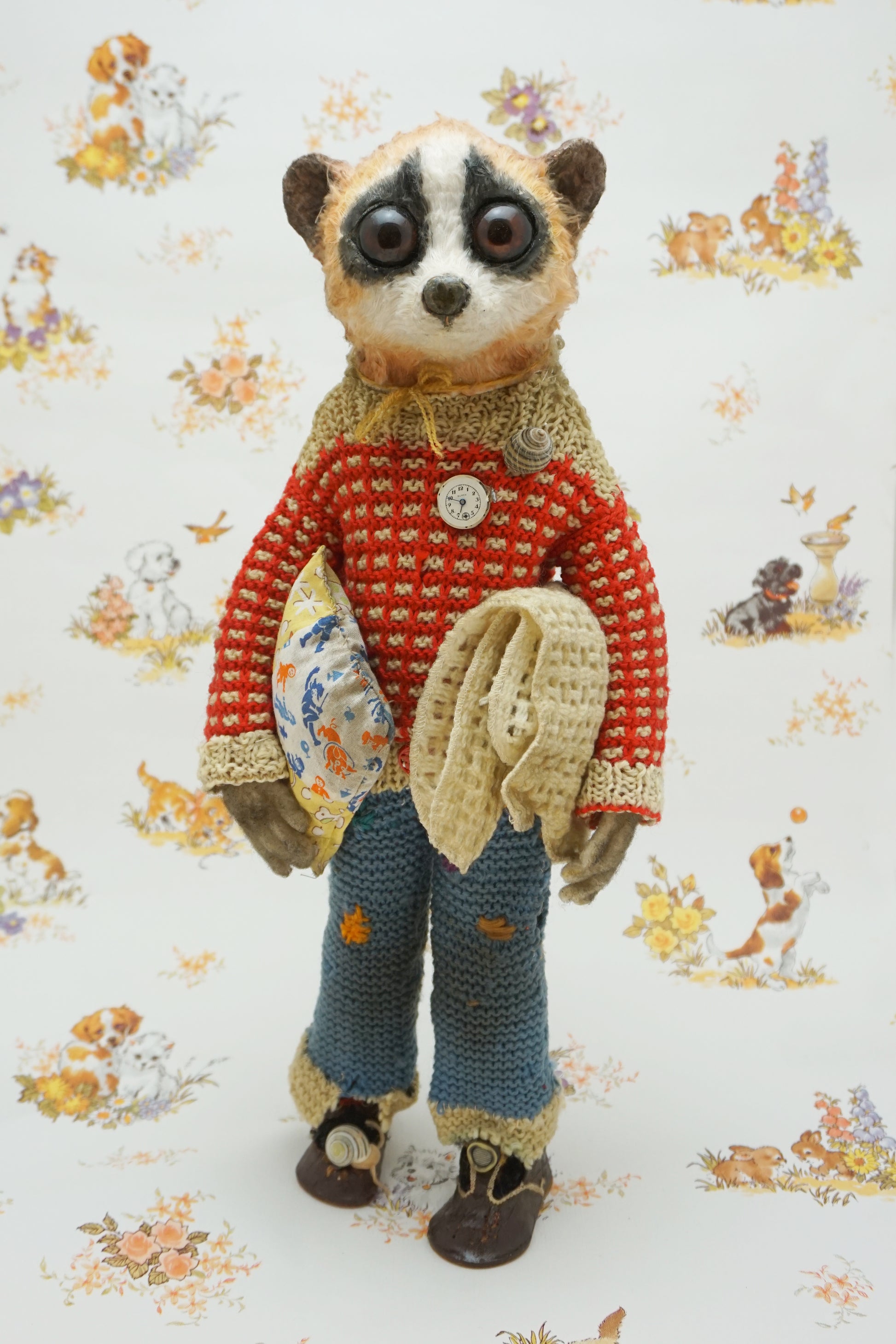 A handmade plush toy of a slow loris dressed in a red knit sweater, blue knit pants, and brown shoes, standing against a white background with a vintage-style animal print.