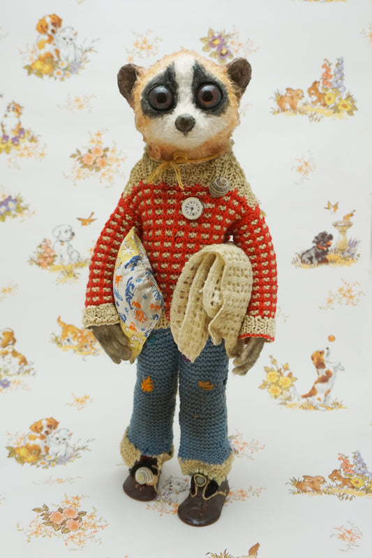 A handmade plush toy of a slow loris dressed in a red knit sweater, blue knit pants, and brown shoes, standing against a white background with a vintage-style animal print.