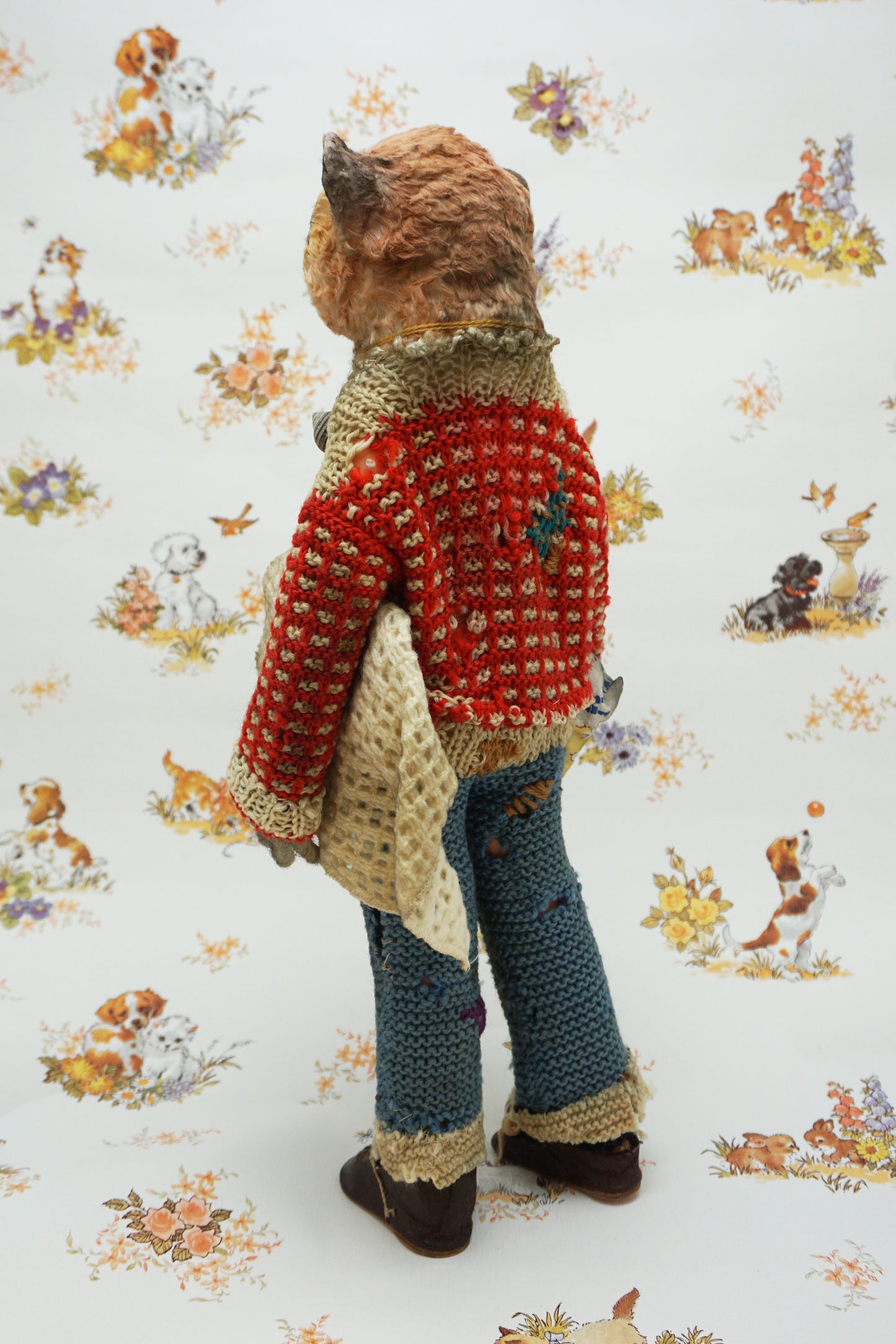 A handmade plush toy of a slow loris dressed in a red knit sweater, blue knit pants, and brown shoes, standing against a white background with a vintage-style animal print.