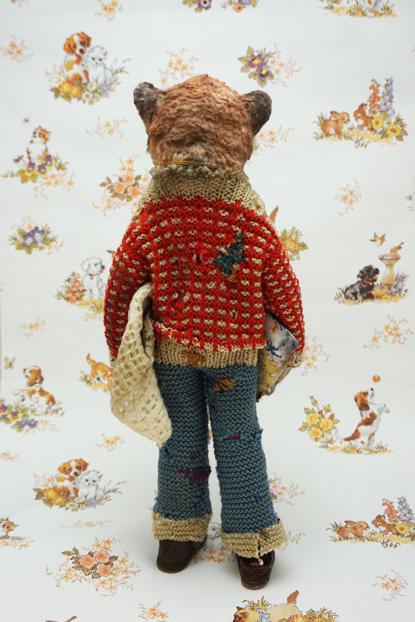 A handmade plush toy of a slow loris dressed in a red knit sweater, blue knit pants, and brown shoes, standing against a white background with a vintage-style animal print.