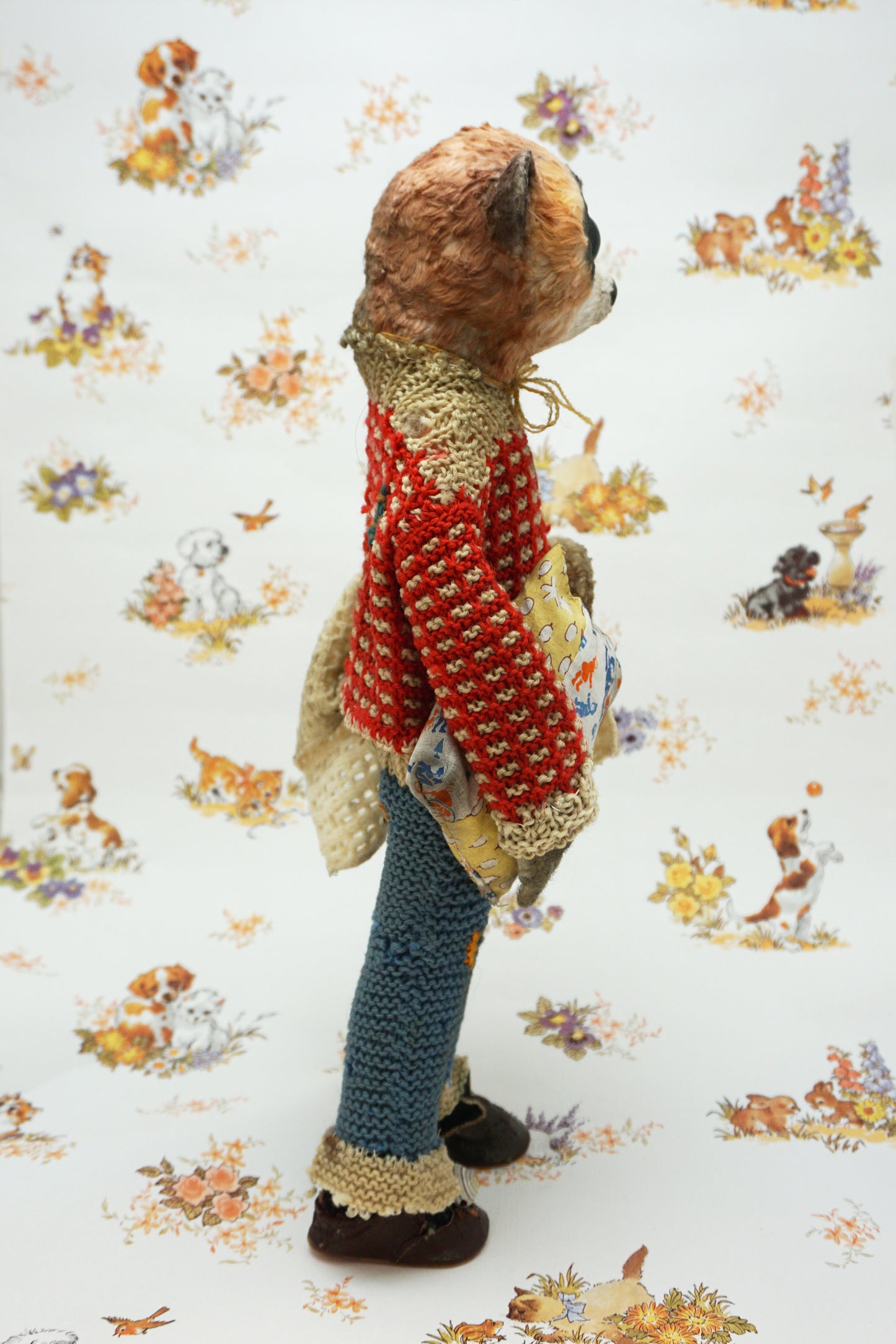 A handmade plush toy of a slow loris dressed in a red knit sweater, blue knit pants, and brown shoes, standing against a white background with a vintage-style animal print.