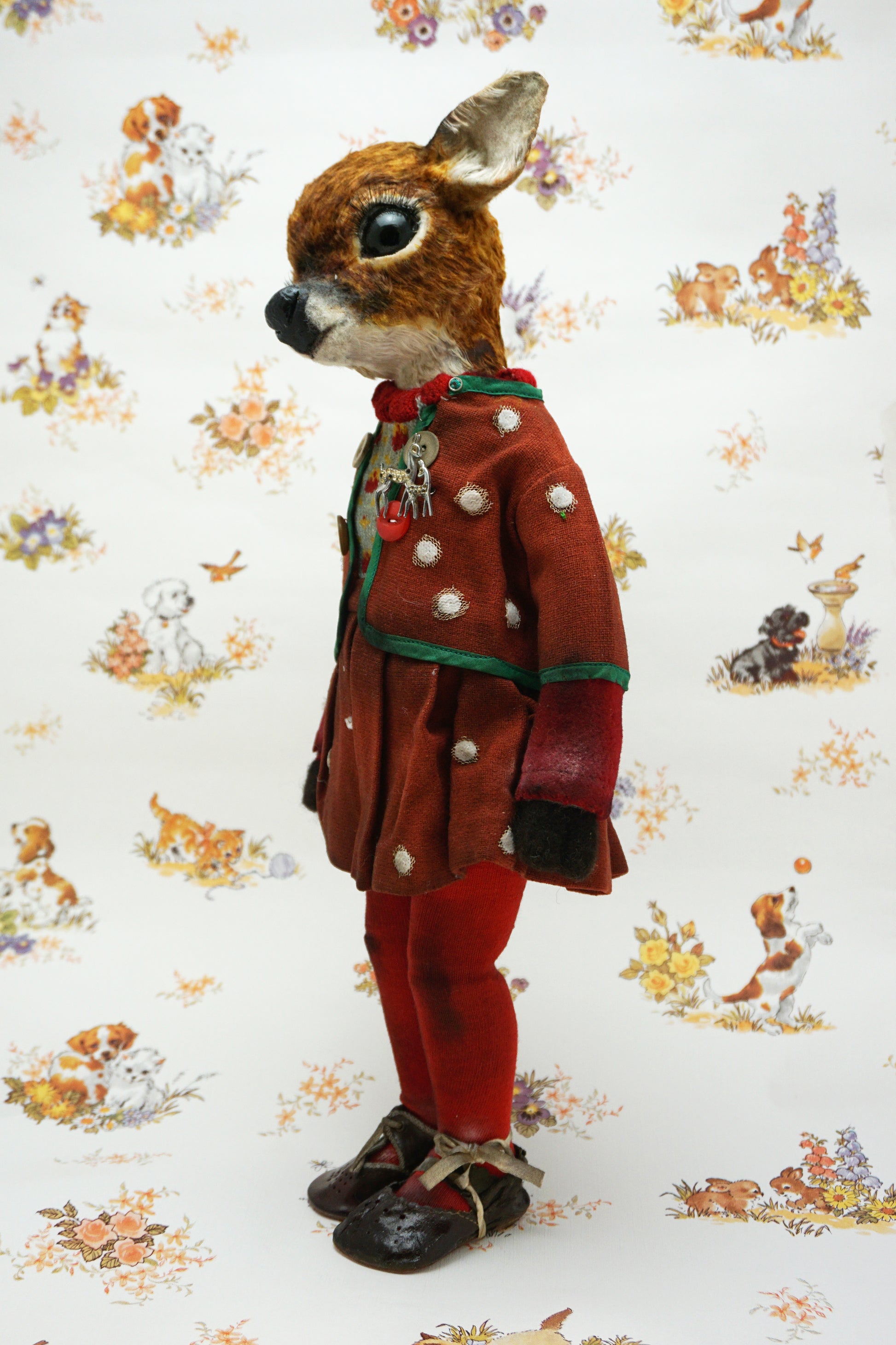 A handmade animal doll with a brown deer head and large dark eyes, dressed in a rust-red velvet jacket with green trim, a knit sweater, red tights, and dark shoes, standing against a white patterned background.
