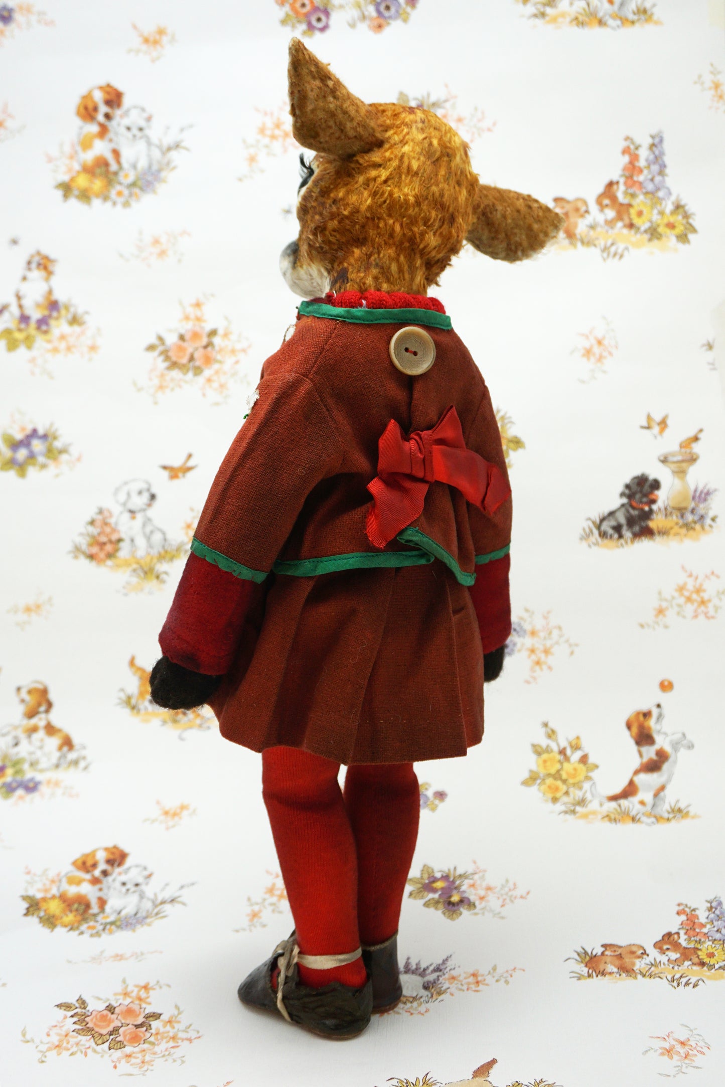 A handmade animal doll with a brown deer head and large dark eyes, dressed in a rust-red velvet jacket with green trim, a knit sweater, red tights, and dark shoes, standing against a white patterned background.
