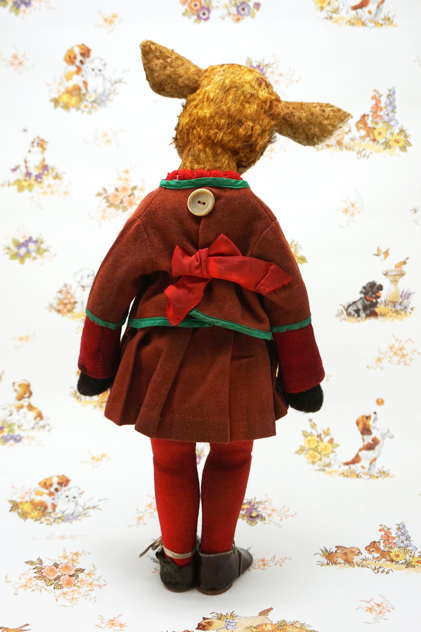 A handmade animal doll with a brown deer head and large dark eyes, dressed in a rust-red velvet jacket with green trim, a knit sweater, red tights, and dark shoes, standing against a white patterned background.