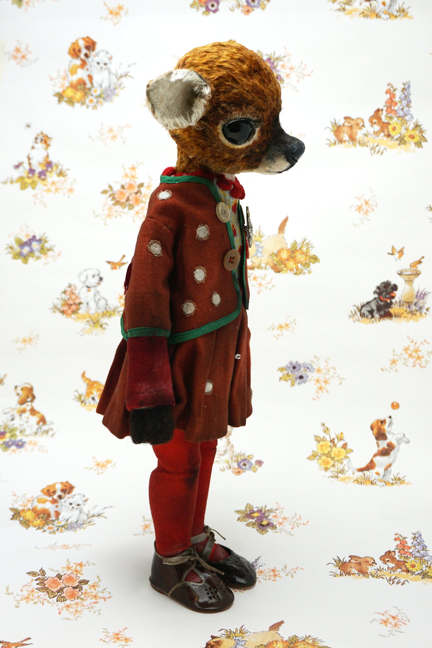 A handmade animal doll with a brown deer head and large dark eyes, dressed in a rust-red velvet jacket with green trim, a knit sweater, red tights, and dark shoes, standing against a white patterned background.