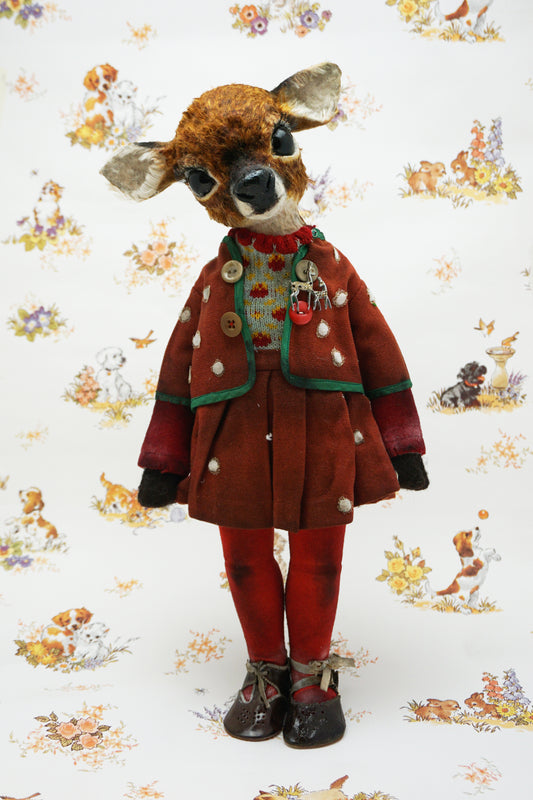 A handmade animal doll with a brown deer head and large dark eyes, dressed in a rust-red velvet jacket with green trim, a knit sweater, red tights, and dark shoes, standing against a white patterned background.
