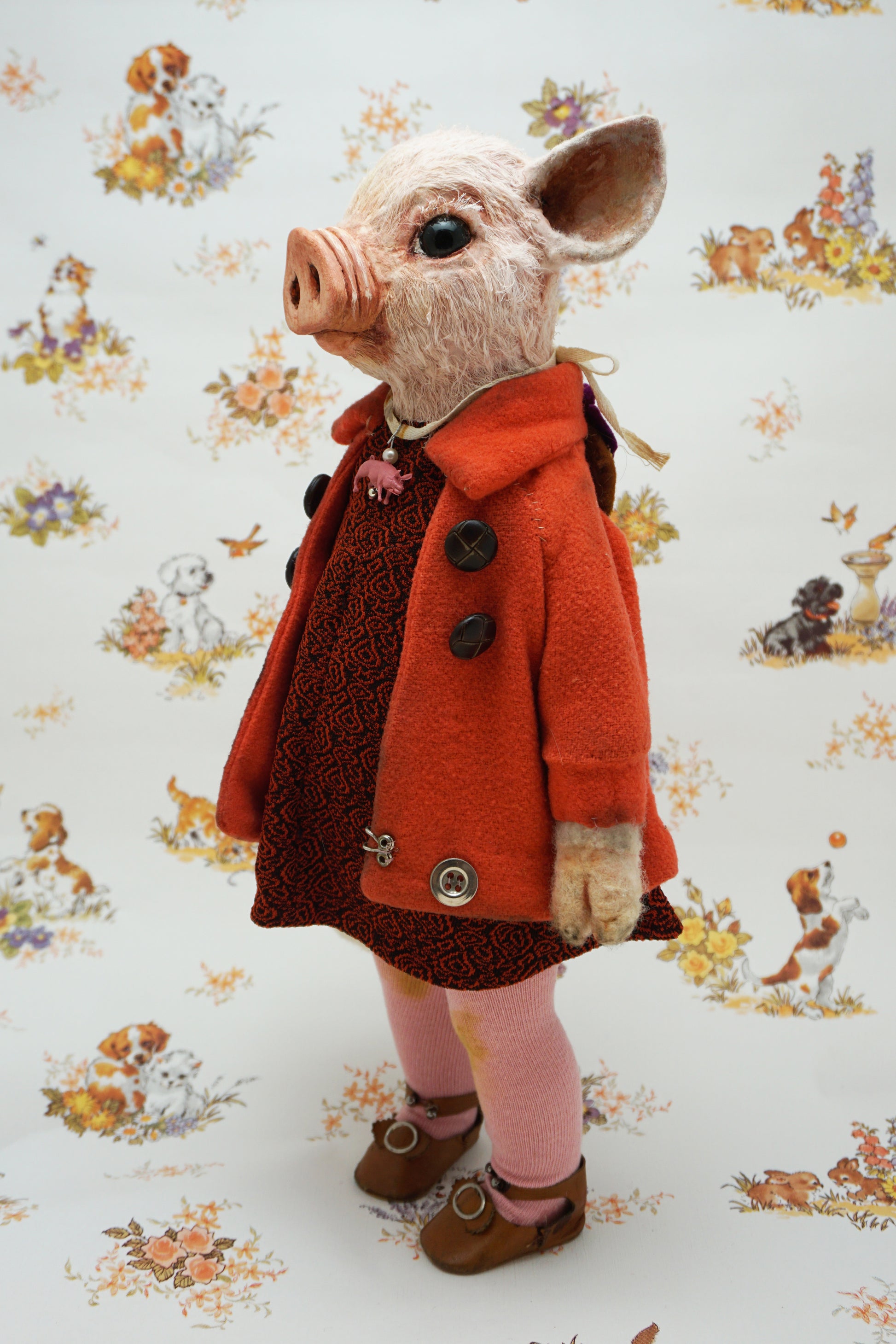 A pig with a pink snout and ears, wearing a bright orange coat over a dark patterned dress, pink tights, and brown shoes, standing against a white patterned backdrop with dog illustrations.