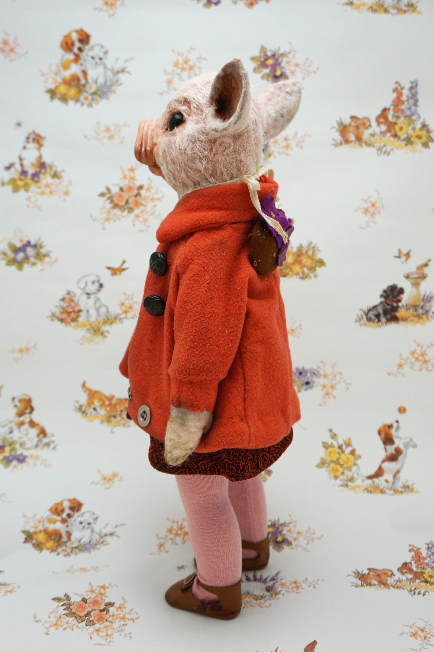 A pig with a pink snout and ears, wearing a bright orange coat over a dark patterned dress, pink tights, and brown shoes, standing against a white patterned backdrop with dog illustrations.