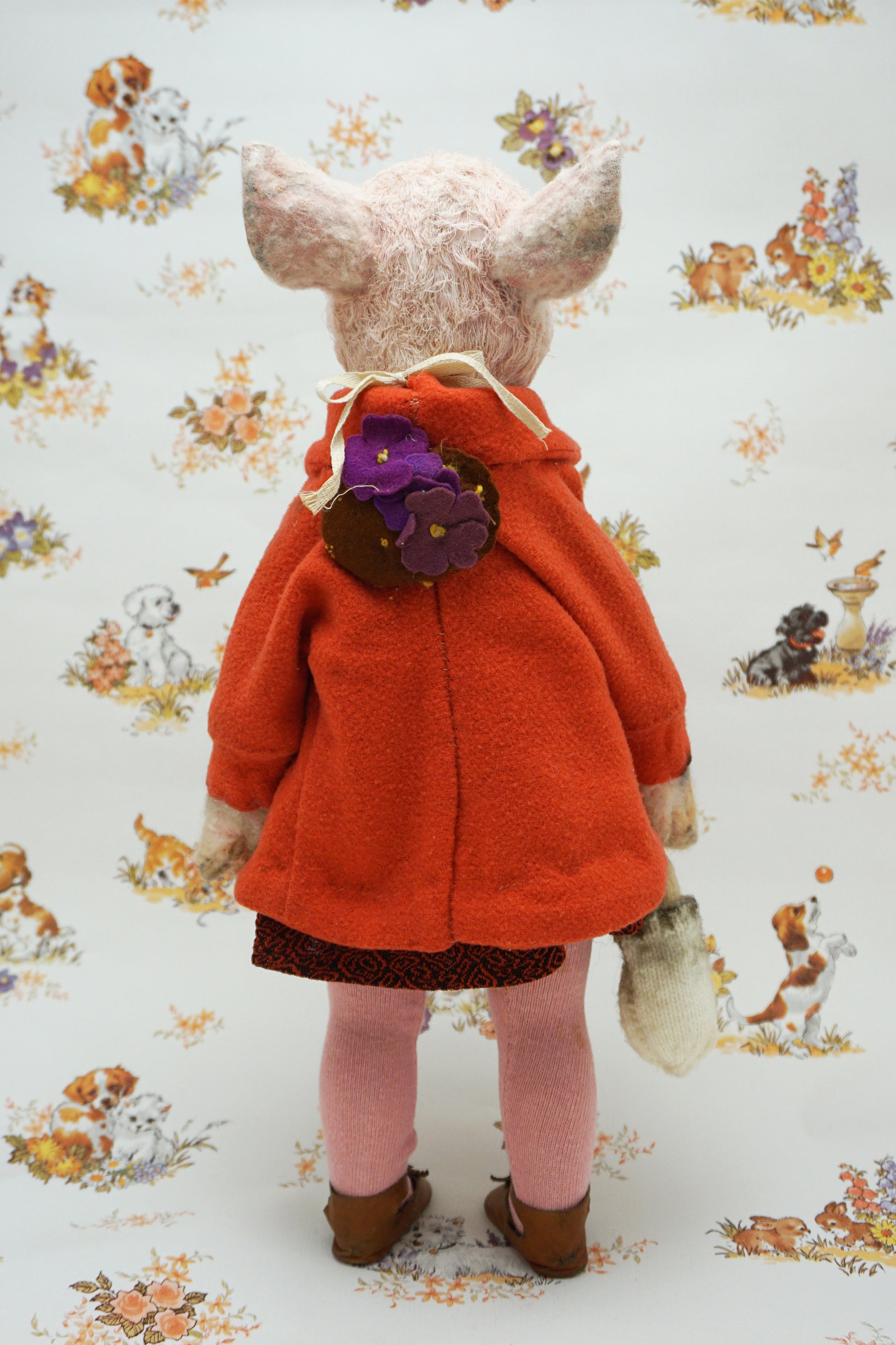 A pig with a pink snout and ears, wearing a bright orange coat over a dark patterned dress, pink tights, and brown shoes, standing against a white patterned backdrop with dog illustrations.