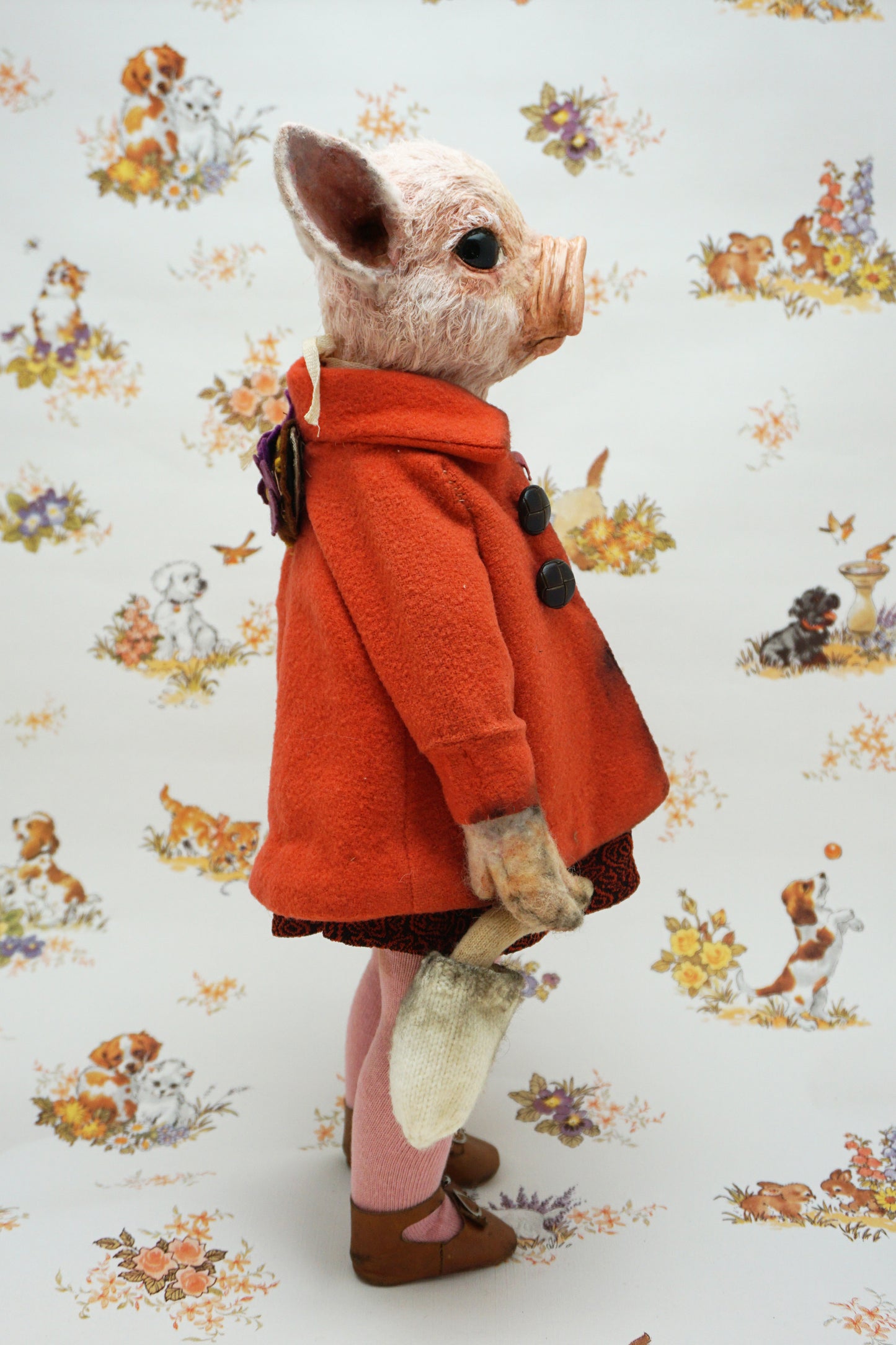 A pig with a pink snout and ears, wearing a bright orange coat over a dark patterned dress, pink tights, and brown shoes, standing against a white patterned backdrop with dog illustrations.