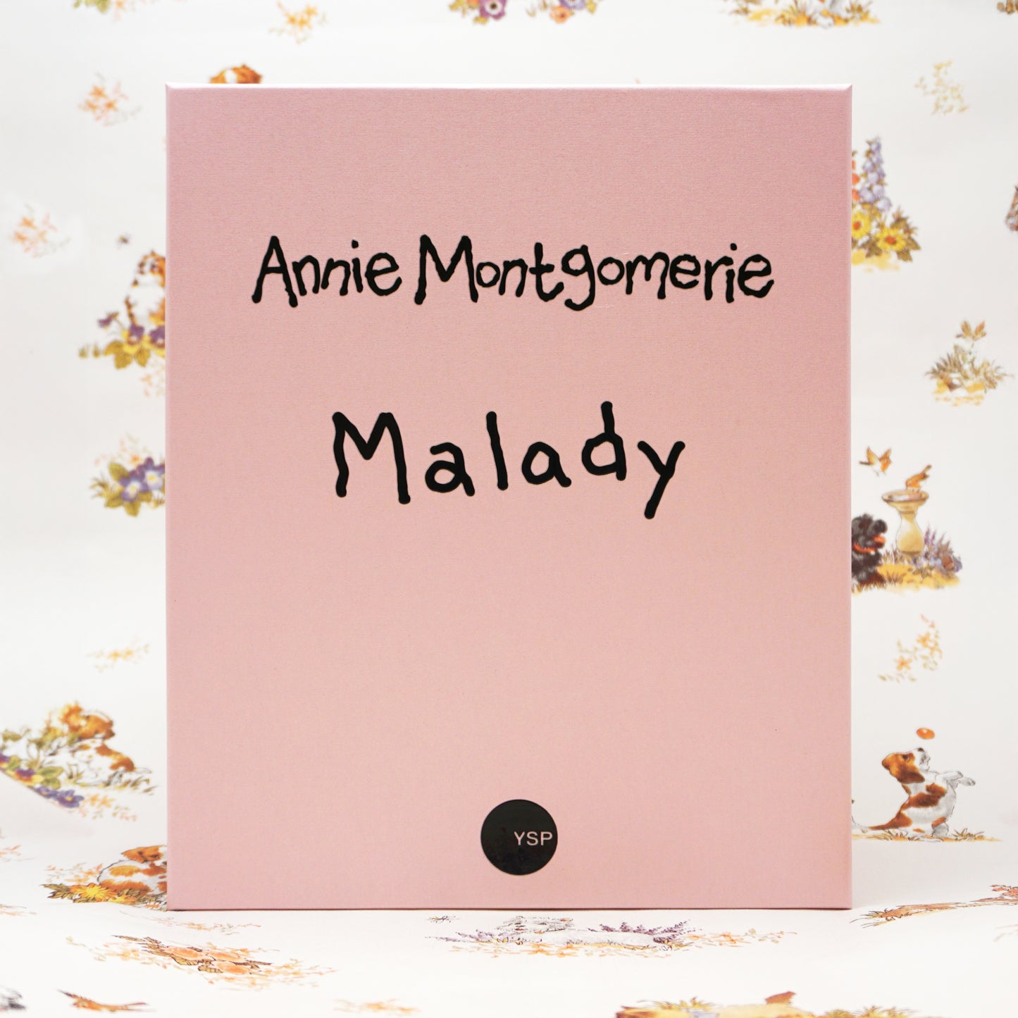 A pink rectangular box standing upright, with the text 'Annie Montgomerie Malady' in black script and a small black circular 'YSP' mark near the bottom. The background is a white floral print surface.