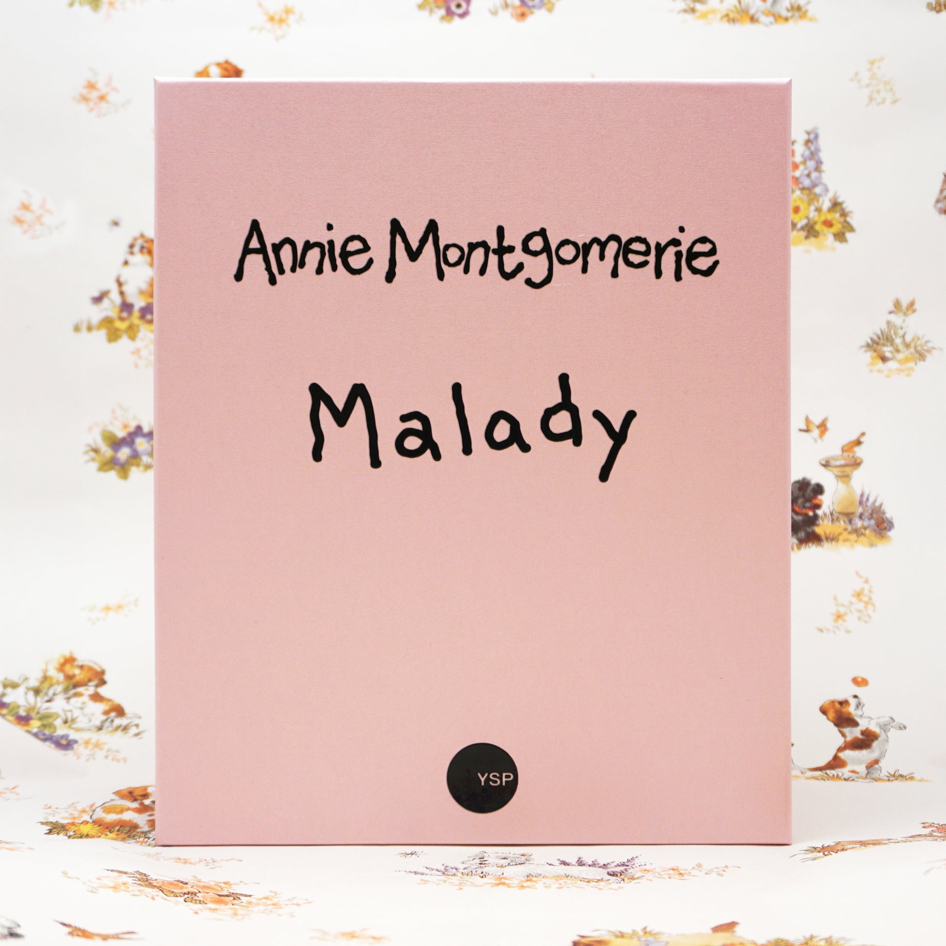 A pink rectangular box standing upright, with the text 'Annie Montgomerie Malady' in black script and a small black circular 'YSP' mark near the bottom. The background is a white floral print surface.