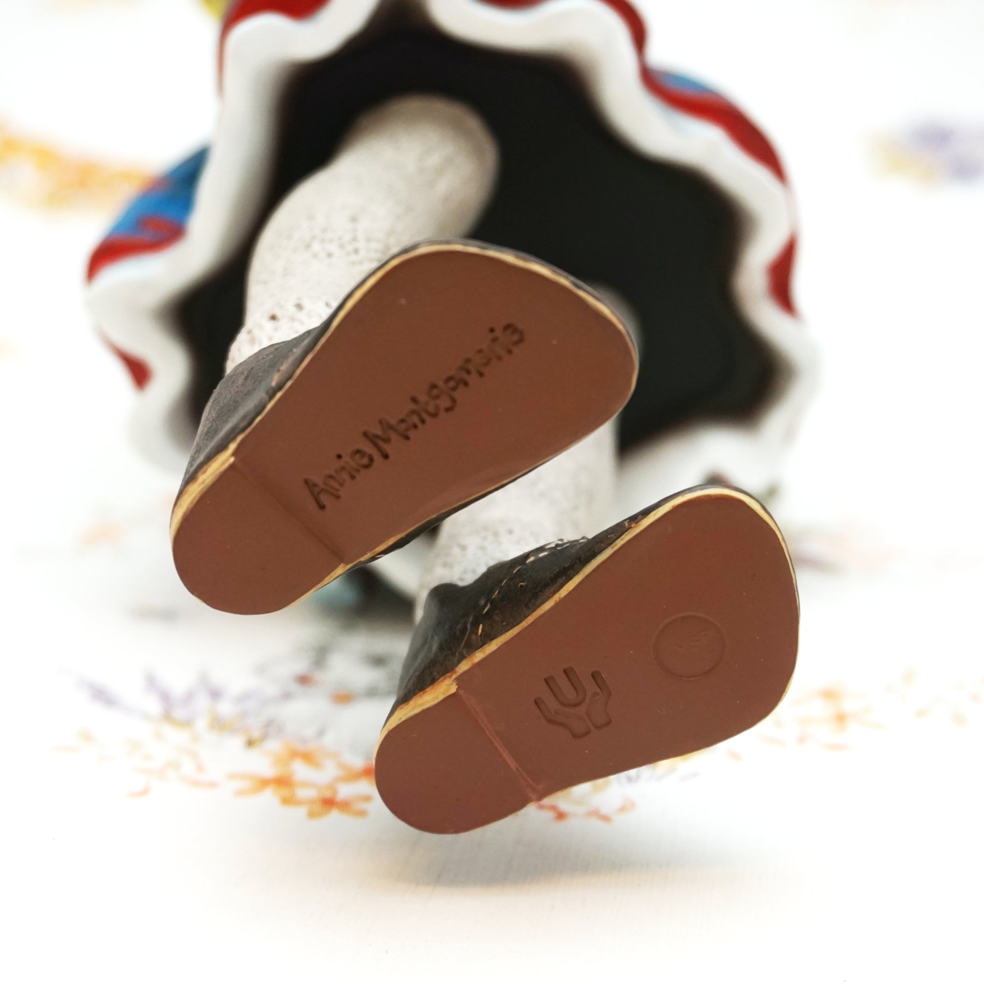 Close-up of the soles of a pair of black shoes with brown outsoles, displayed on a white surface with a colourful ruffled fabric in the background. The name 'Annie Montgomerie' is embossed on the left sole.