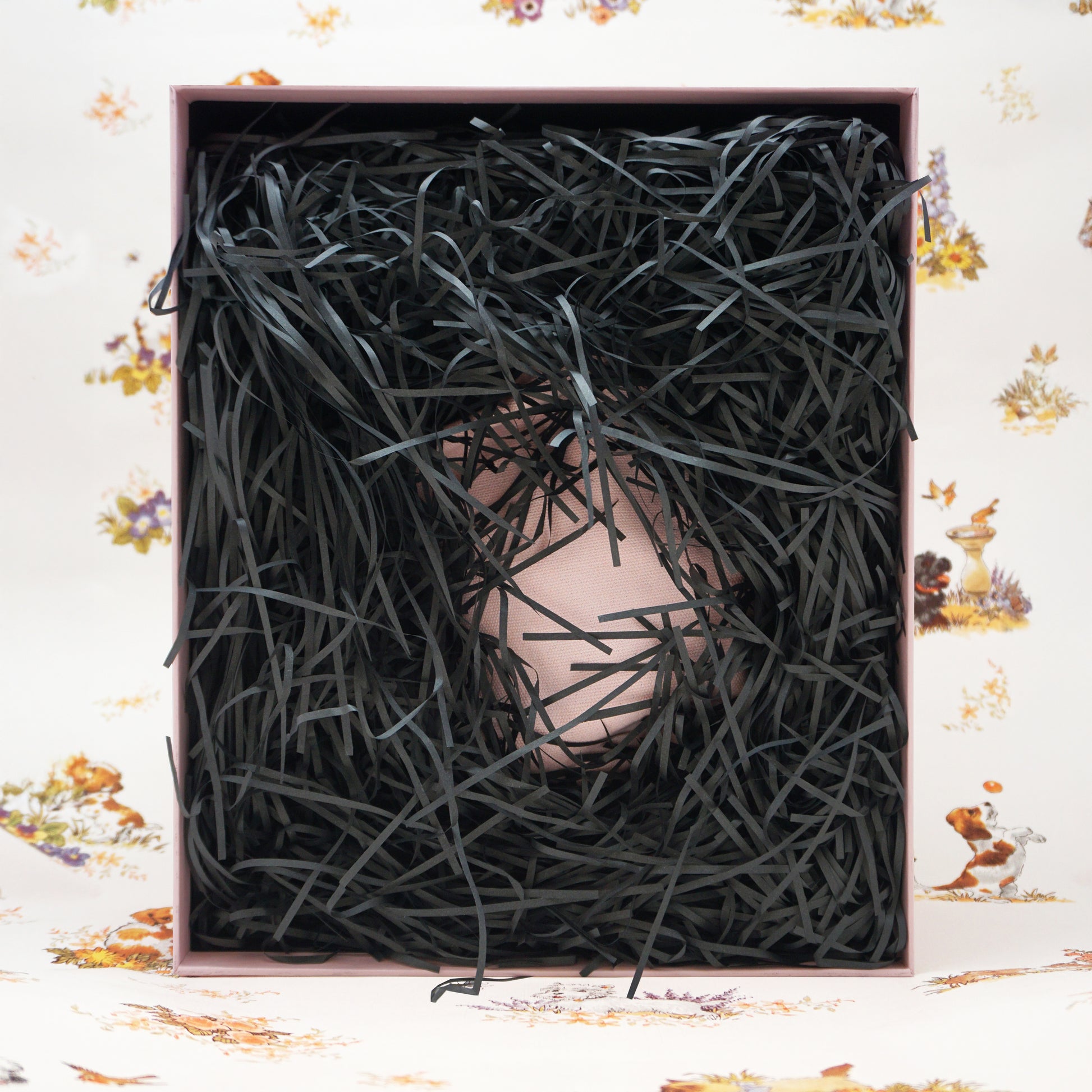 A pale pink drawstring bag nestled inside a square box filled with black shredded paper crinkle packing, set against a white floral background.