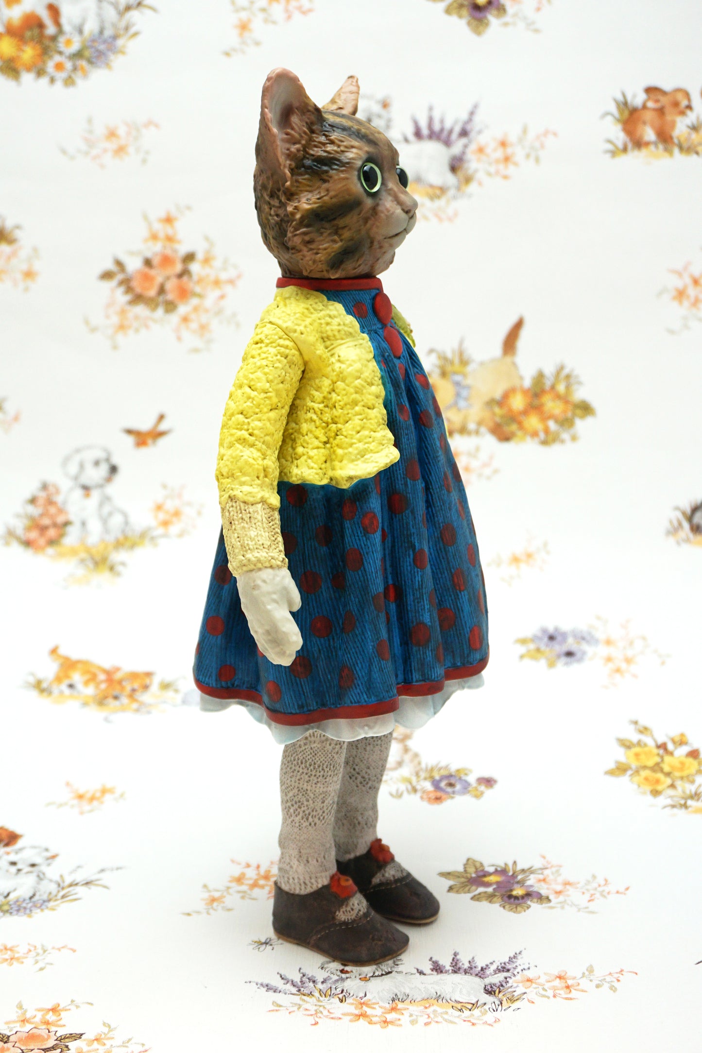 A collectable vinyl figure of a cat dressed in a handmade outfit, featuring a yellow knit cardigan, a blue polka-dot dress, beige socks, and brown shoes, standing against a white floral-patterned backdrop.