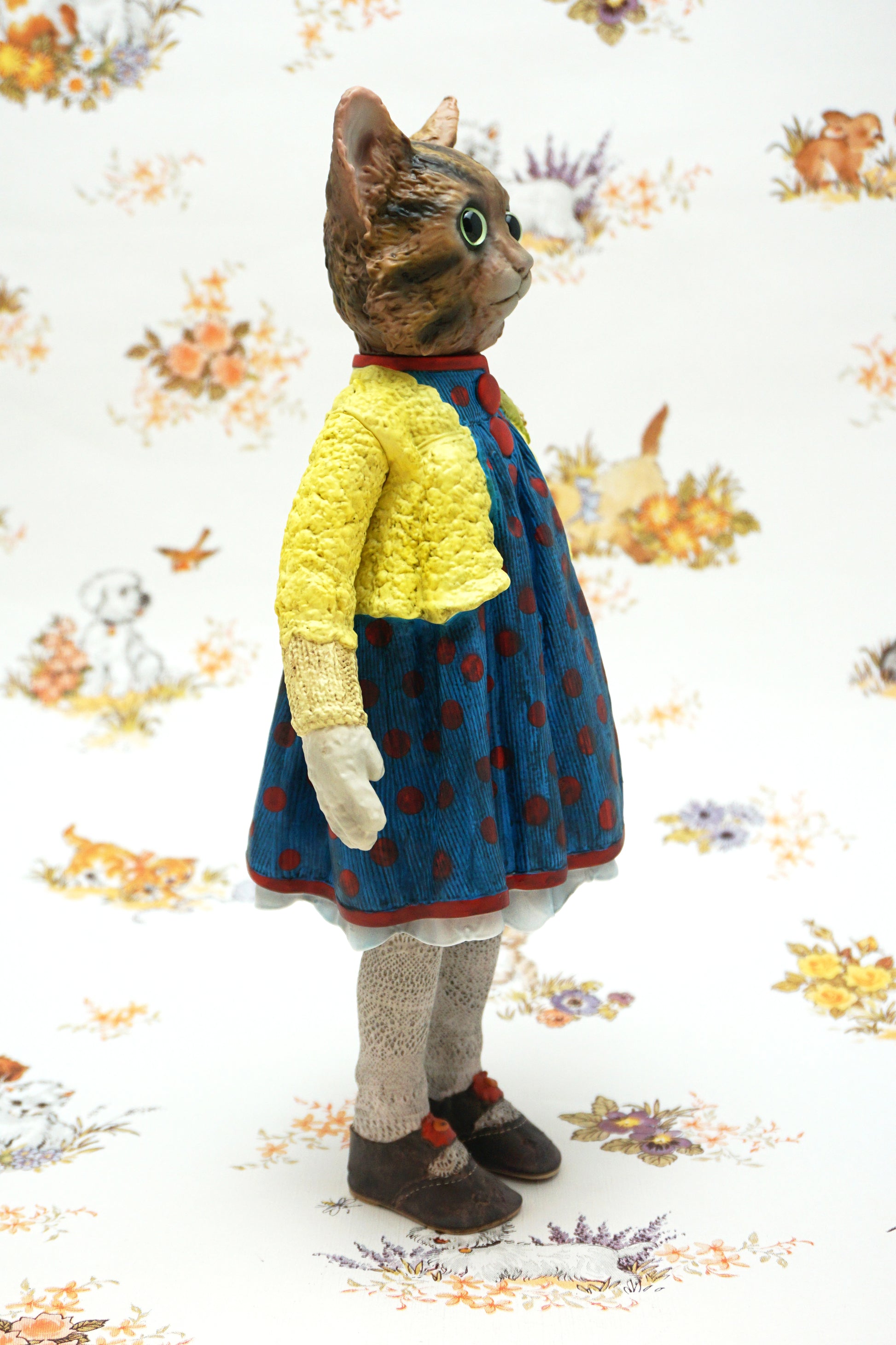 A collectable vinyl figure of a cat dressed in a handmade outfit, featuring a yellow knit cardigan, a blue polka-dot dress, beige socks, and brown shoes, standing against a white floral-patterned backdrop.
