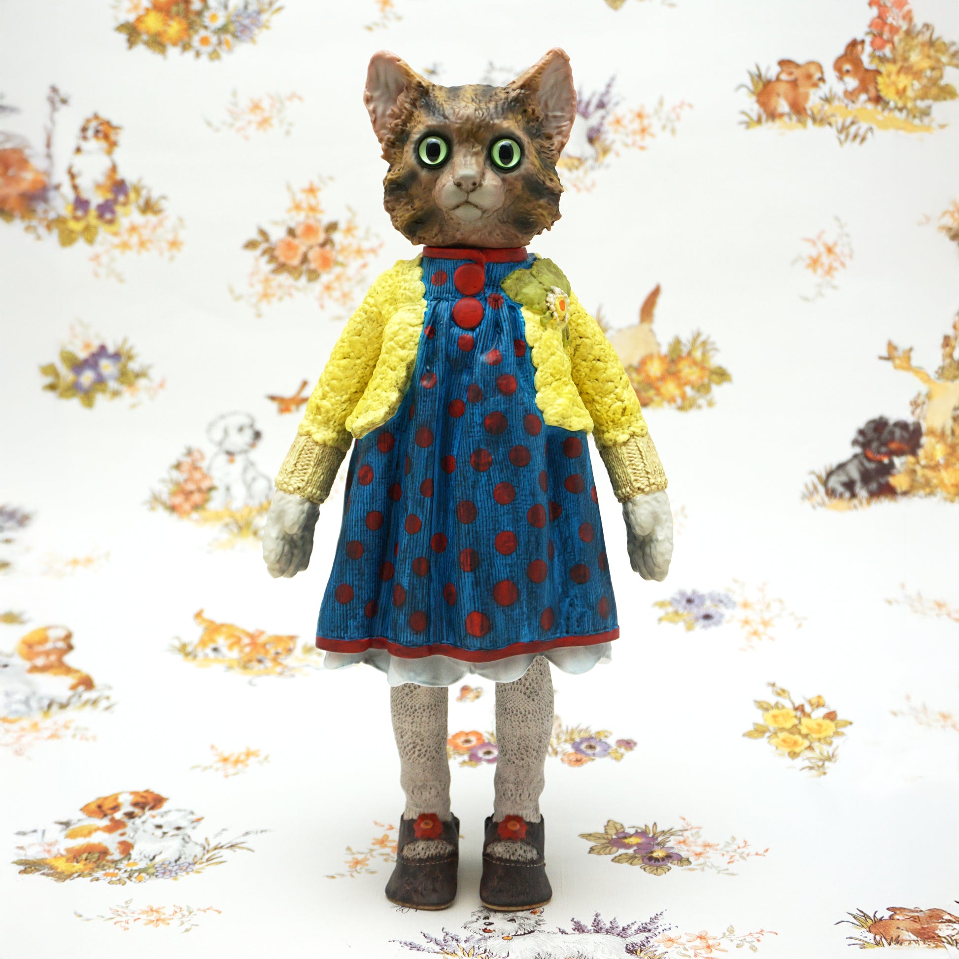 A highly detailed collectible figurine of a cat dressed in a blue polka-dot dress and yellow cardigan, standing against a patterned white background.