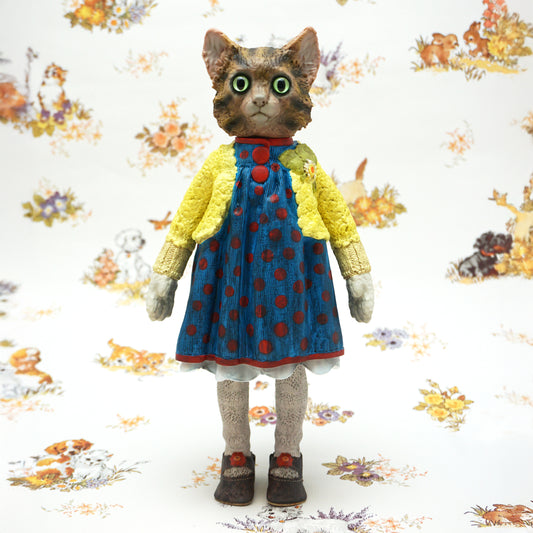 A highly detailed collectible figurine of a cat dressed in a blue polka-dot dress and yellow cardigan, standing against a patterned white background.