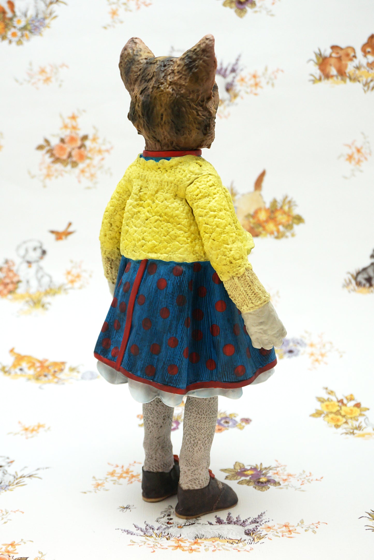 A collectable vinyl figure of a cat dressed in a handmade outfit, featuring a yellow knit cardigan, a blue polka-dot dress, beige socks, and brown shoes, standing against a white floral-patterned backdrop.
