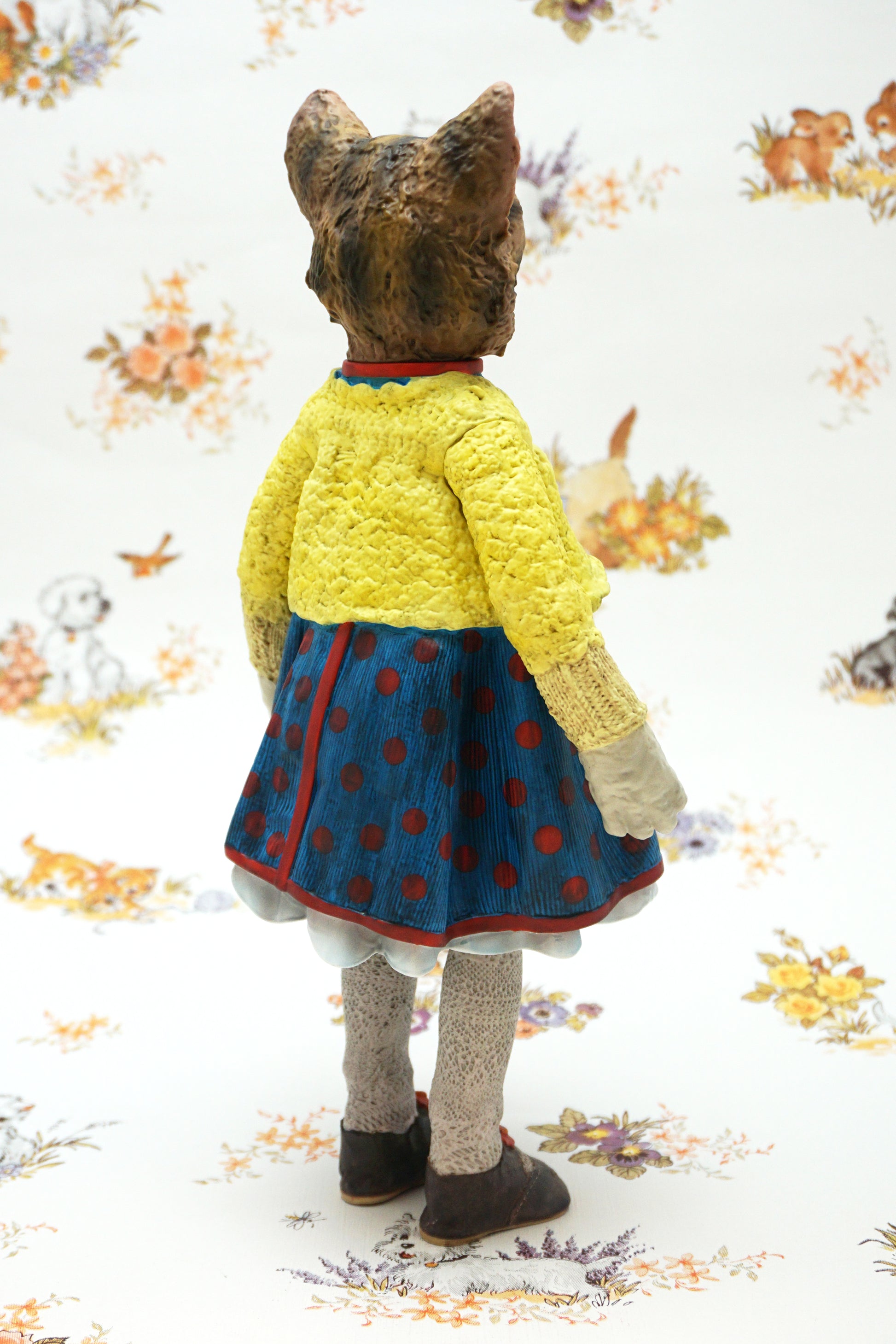 A collectable vinyl figure of a cat dressed in a handmade outfit, featuring a yellow knit cardigan, a blue polka-dot dress, beige socks, and brown shoes, standing against a white floral-patterned backdrop.