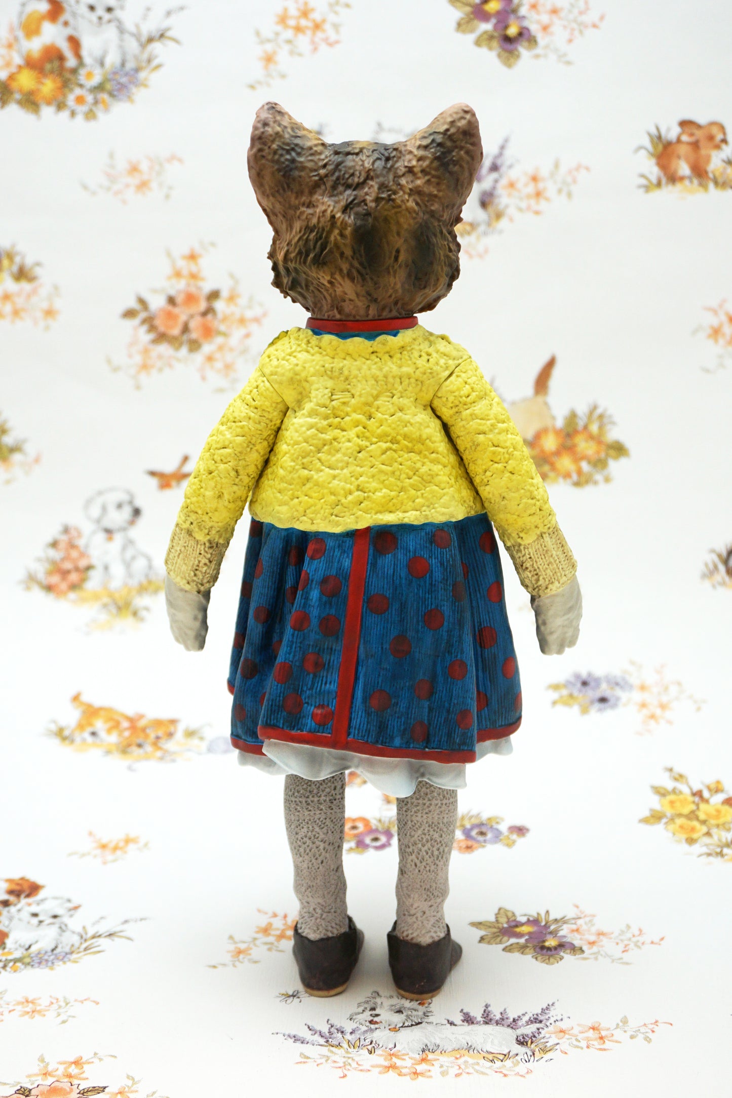 A collectable vinyl figure of a cat dressed in a handmade outfit, featuring a yellow knit cardigan, a blue polka-dot dress, beige socks, and brown shoes, standing against a white floral-patterned backdrop.