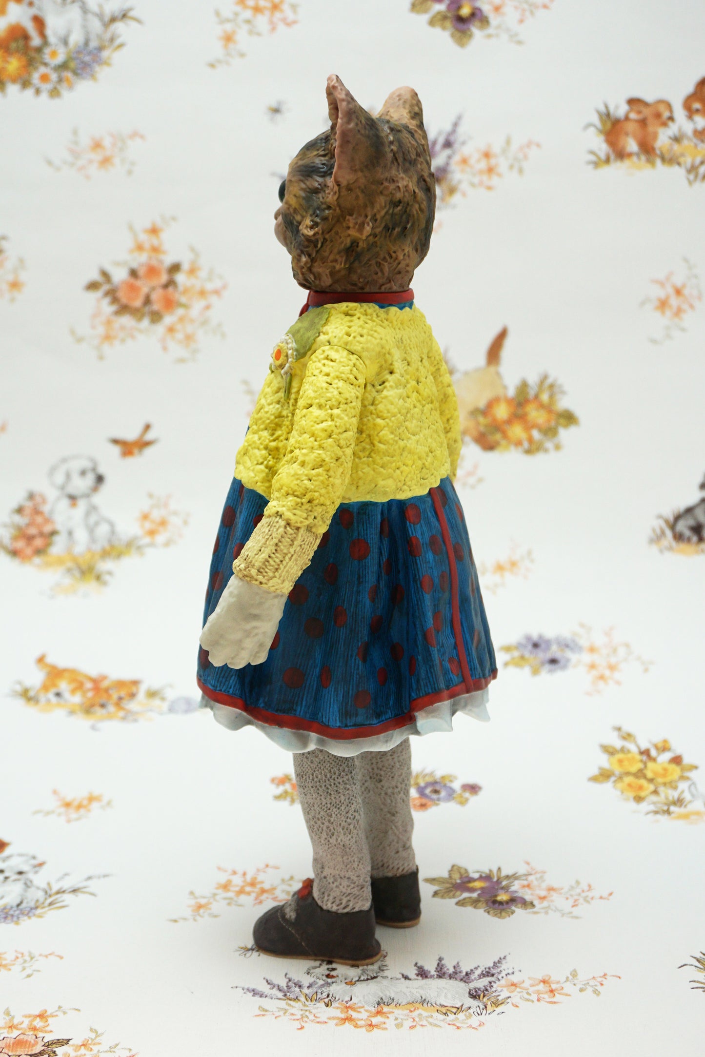 A collectable vinyl figure of a cat dressed in a handmade outfit, featuring a yellow knit cardigan, a blue polka-dot dress, beige socks, and brown shoes, standing against a white floral-patterned backdrop.
