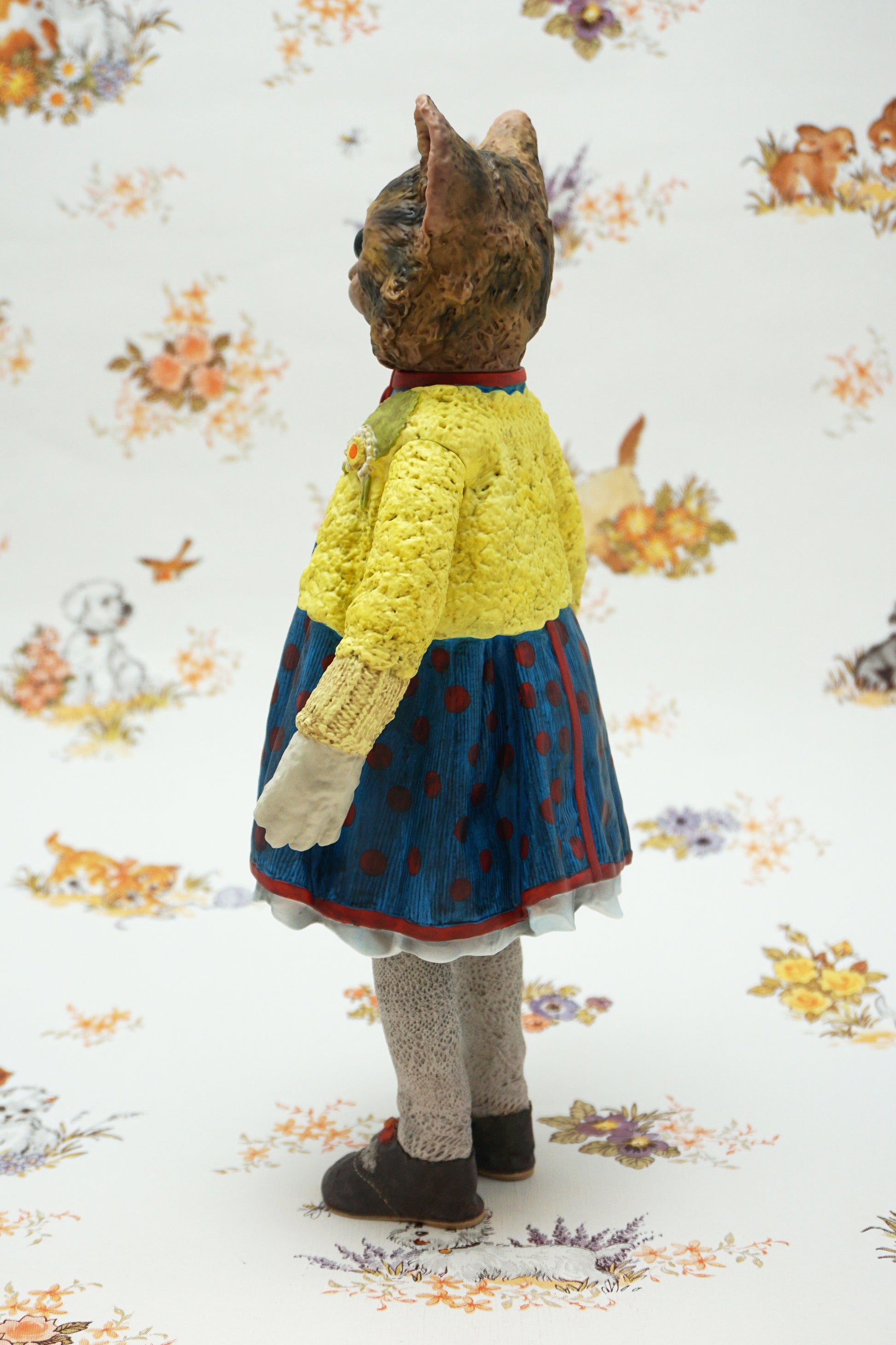 A collectable vinyl figure of a cat dressed in a handmade outfit, featuring a yellow knit cardigan, a blue polka-dot dress, beige socks, and brown shoes, standing against a white floral-patterned backdrop.