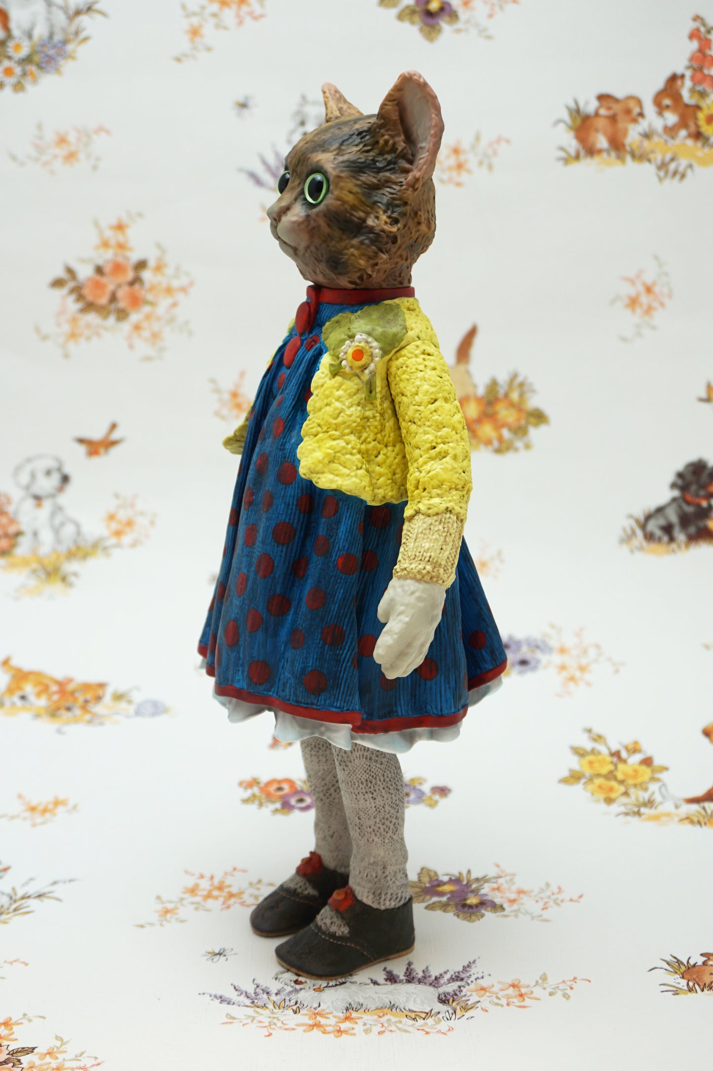 A collectable vinyl figure of a cat dressed in a handmade outfit, featuring a yellow knit cardigan, a blue polka-dot dress, beige socks, and brown shoes, standing against a white floral-patterned backdrop.