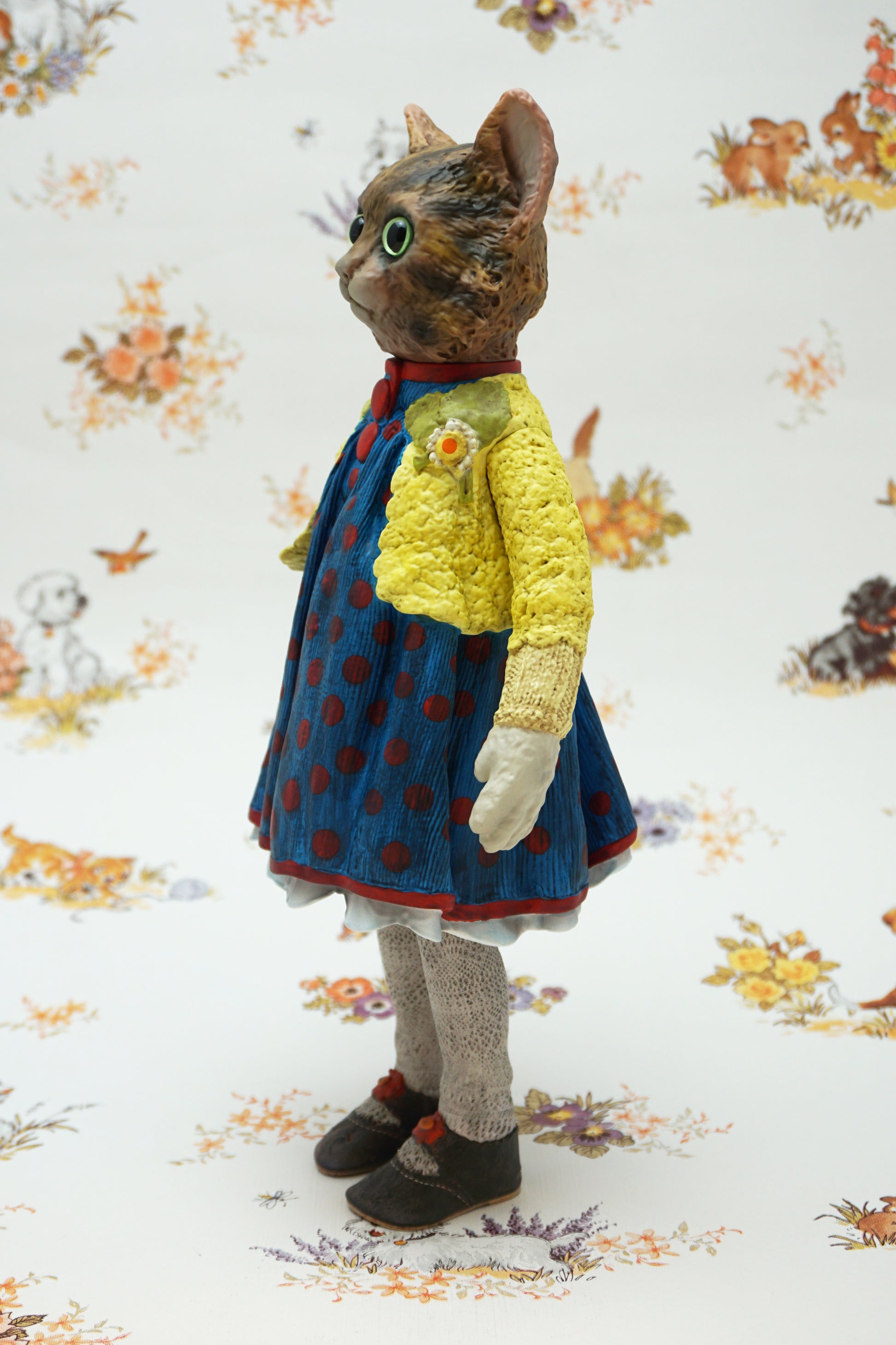 A collectable vinyl figure of a cat dressed in a handmade outfit, featuring a yellow knit cardigan, a blue polka-dot dress, beige socks, and brown shoes, standing against a white floral-patterned backdrop.