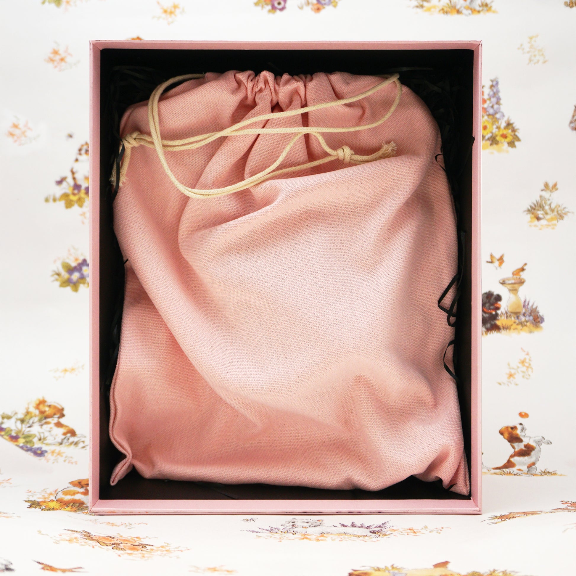 A soft blush pink drawstring bag inside an open pink gift box, set against a white floral background.
