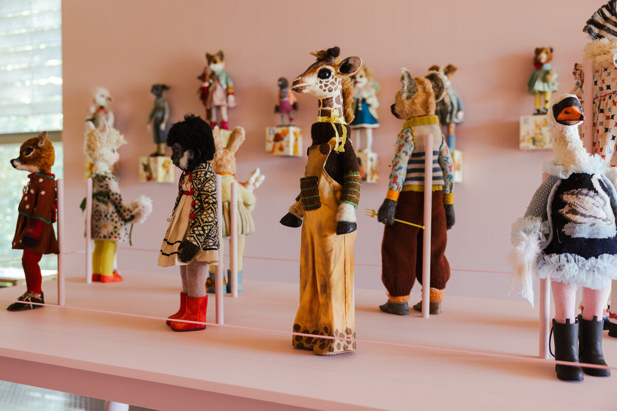 A gallery display featuring a collection of colourful, hand-crafted animal dolls, each dressed in unique knitted or patterned clothing.